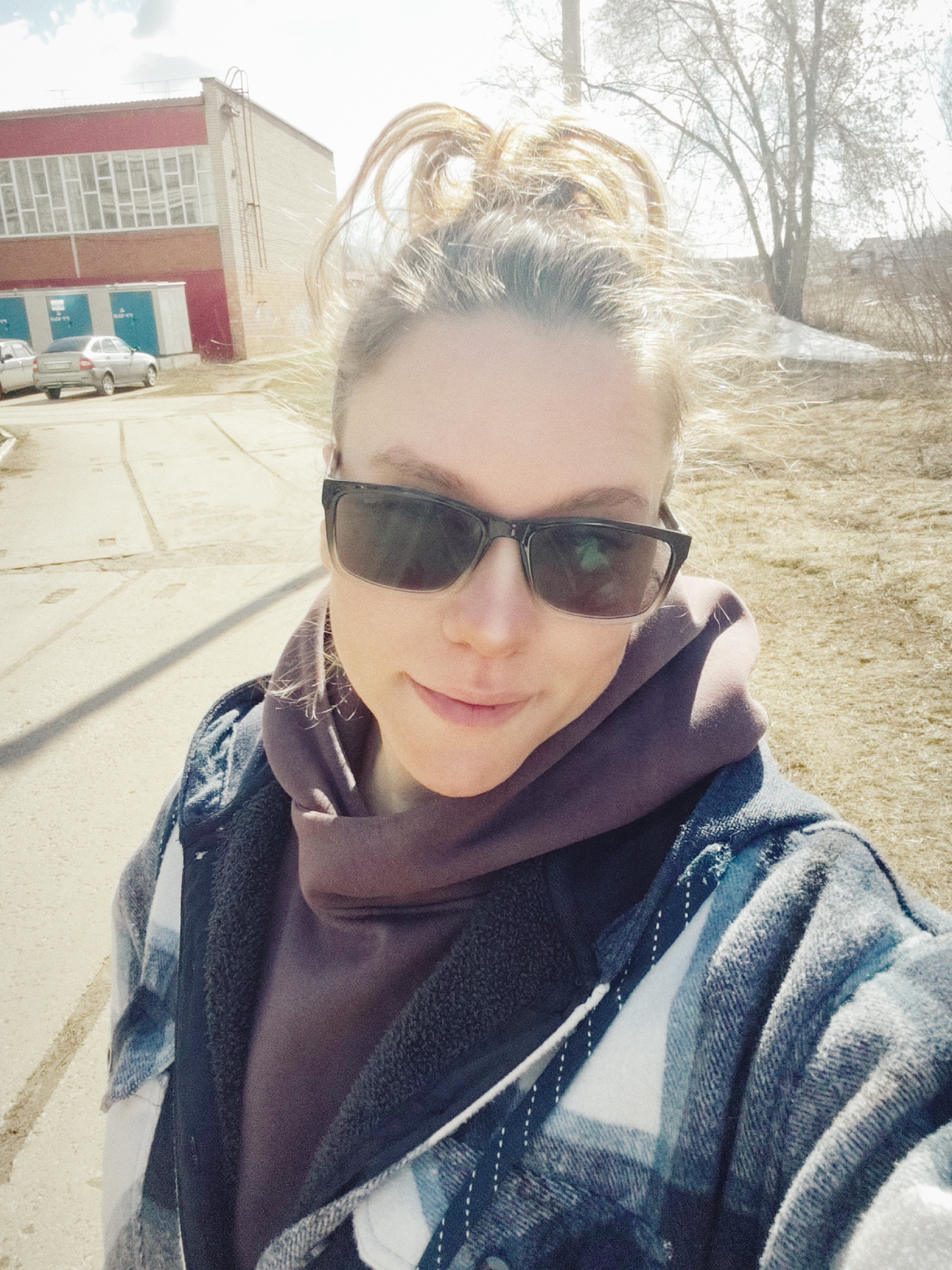 A young woman stands outdoors, taking a selfie. She has her blonde hair pulled up in a messy bun, and she is wearing sunglasses, a brown hoodie, and a blue and white plaid shirt. The background shows an urban environment, possibly the city of Chaikovsky, Russia, based on the presence of a utility pole, a red brick building with several windows, a small gray car parked nearby, a patch of ground with dry grass, and bare trees in the distance. A sidewalk is visible in the foreground. It appears to be a sunny day.