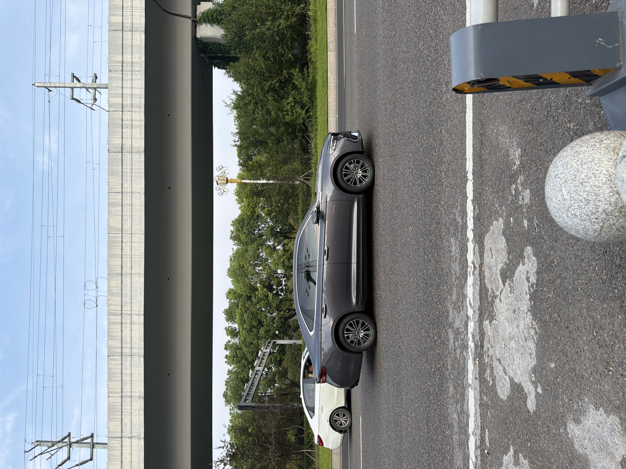 In Jiangjin, China, a grey sedan is driving on an asphalt road, with a white sedan to its left. Overhead, a concrete overpass supports electrical wires and utility poles. The background features a line of green trees and a cloudy sky. A traffic barrier with yellow and black stripes is visible in the foreground on the right side of the image.