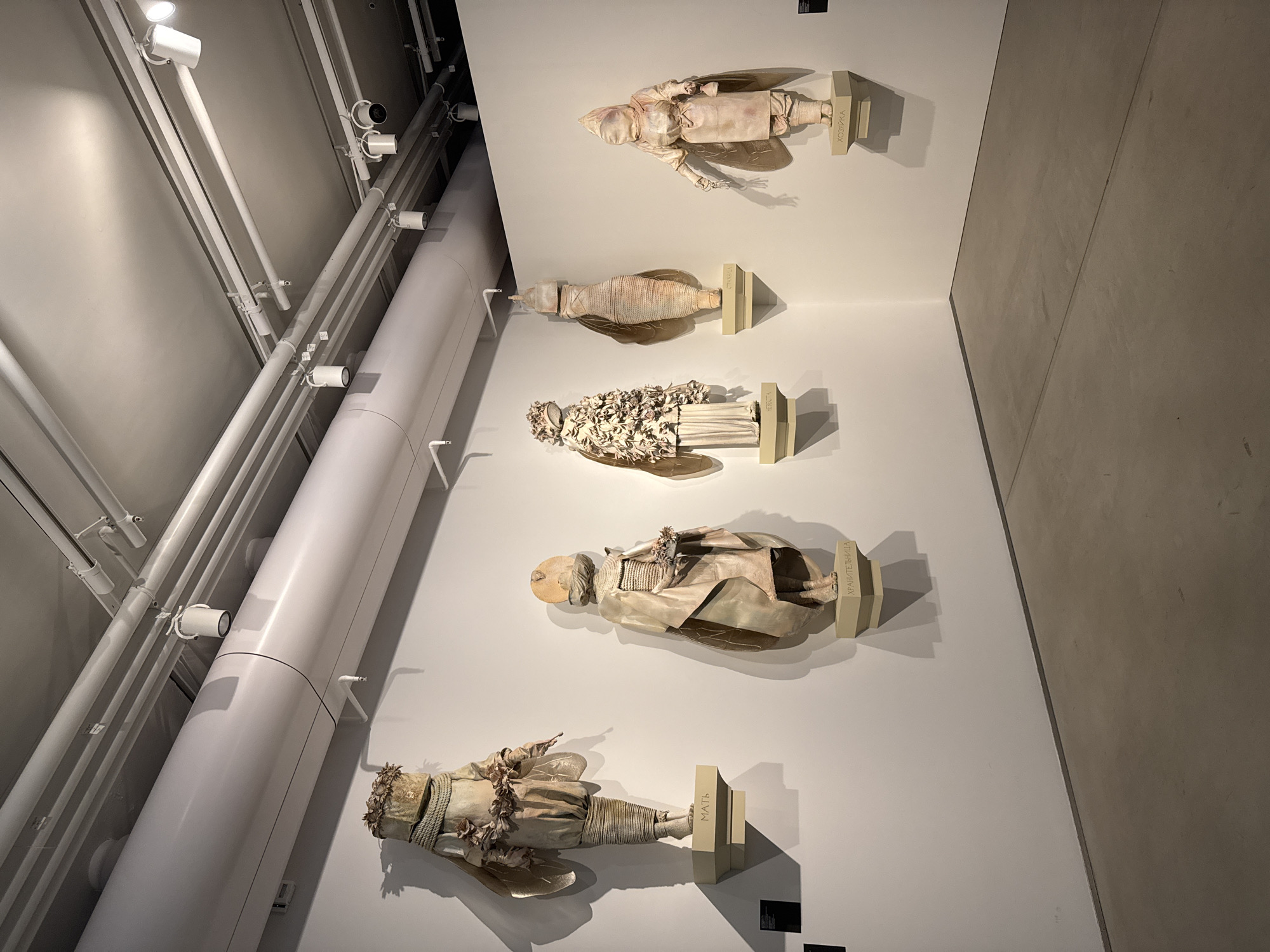 The image shows an art installation featuring five doll-like figures mounted on a white wall. The figures are arranged vertically, each supported by a small stand inscribed with text. The figures vary in design and detail, appearing to be constructed from fabric or other pliable materials. The background consists of a white wall and a complex array of pipes, beams, and lighting fixtures typical of a museum setting. Given the presence of inscriptions in what appears to be Russian and the aesthetic of the art, the location is likely Moscow, Russia.