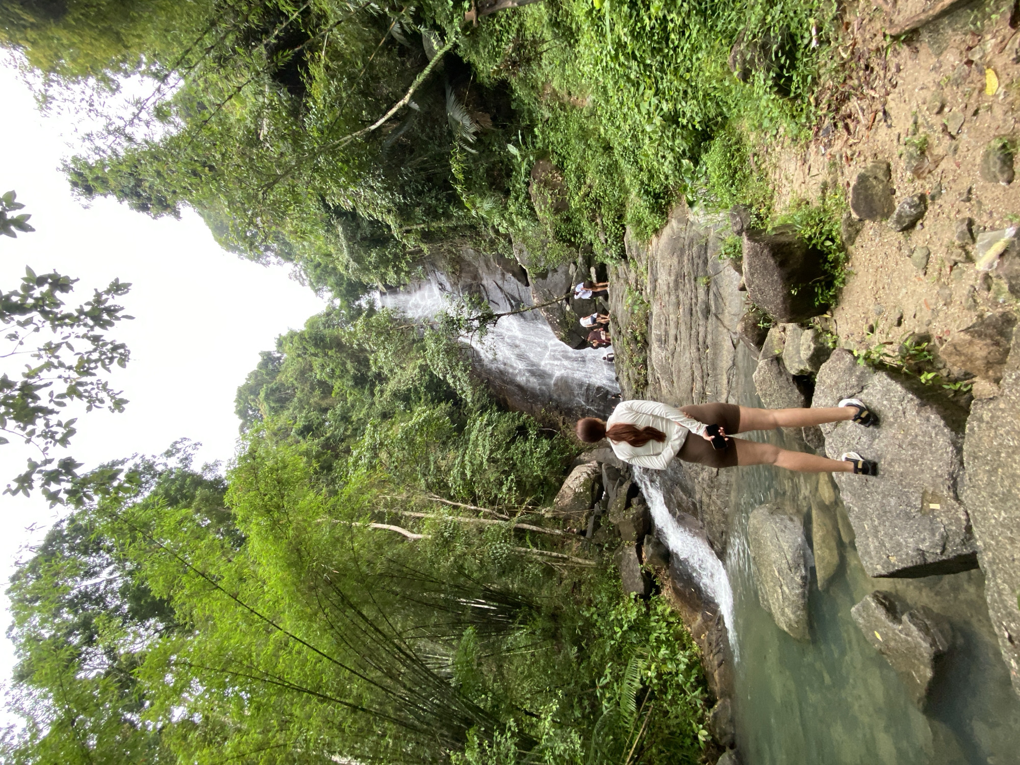 A partially rotated, eye-level photograph captures a waterfall scene in Ban Bang Khli, Thailand. A woman stands on large rocks in the foreground, gazing toward the cascading water. She is wearing a light-colored jacket, brown shorts, and sandals.
The waterfall is surrounded by lush green vegetation. In the background, several people are near the waterfall's higher reaches. Bamboo plants are visible in the lower left of the frame. The water flows into a clear pool. The surrounding environment consists of rocks, foliage, and a dense, green forest.