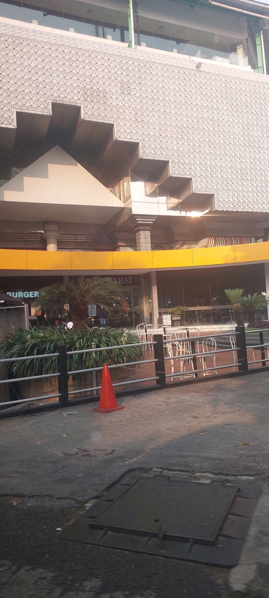 The image captures a street-level view of a modern building with a textured facade, situated in Surabaya, Indonesia. The building's architecture features angular, stepped elements on its upper levels. A bright yellow band runs across the middle section of the building. Below this band, large glass windows reveal the interior of what appears to be a restaurant or cafe, with tables and chairs visible. Signs for "BURGER" and "BARAT" are partially visible on the storefront.
In the foreground, a black metal fence with horizontal railings separates the sidewalk from a planter filled with dense green foliage. A single orange traffic cone stands on the concrete ground in front of the fence. The concrete surface in the foreground shows signs of wear, with cracks and a dark, square manhole cover. The lighting suggests it is either early morning or late afternoon, with long shadows cast across the ground. Several people are visible inside the establishment, seemingly engaged in dining or conversation. The overall scene depicts an urban commercial area in Surabaya.