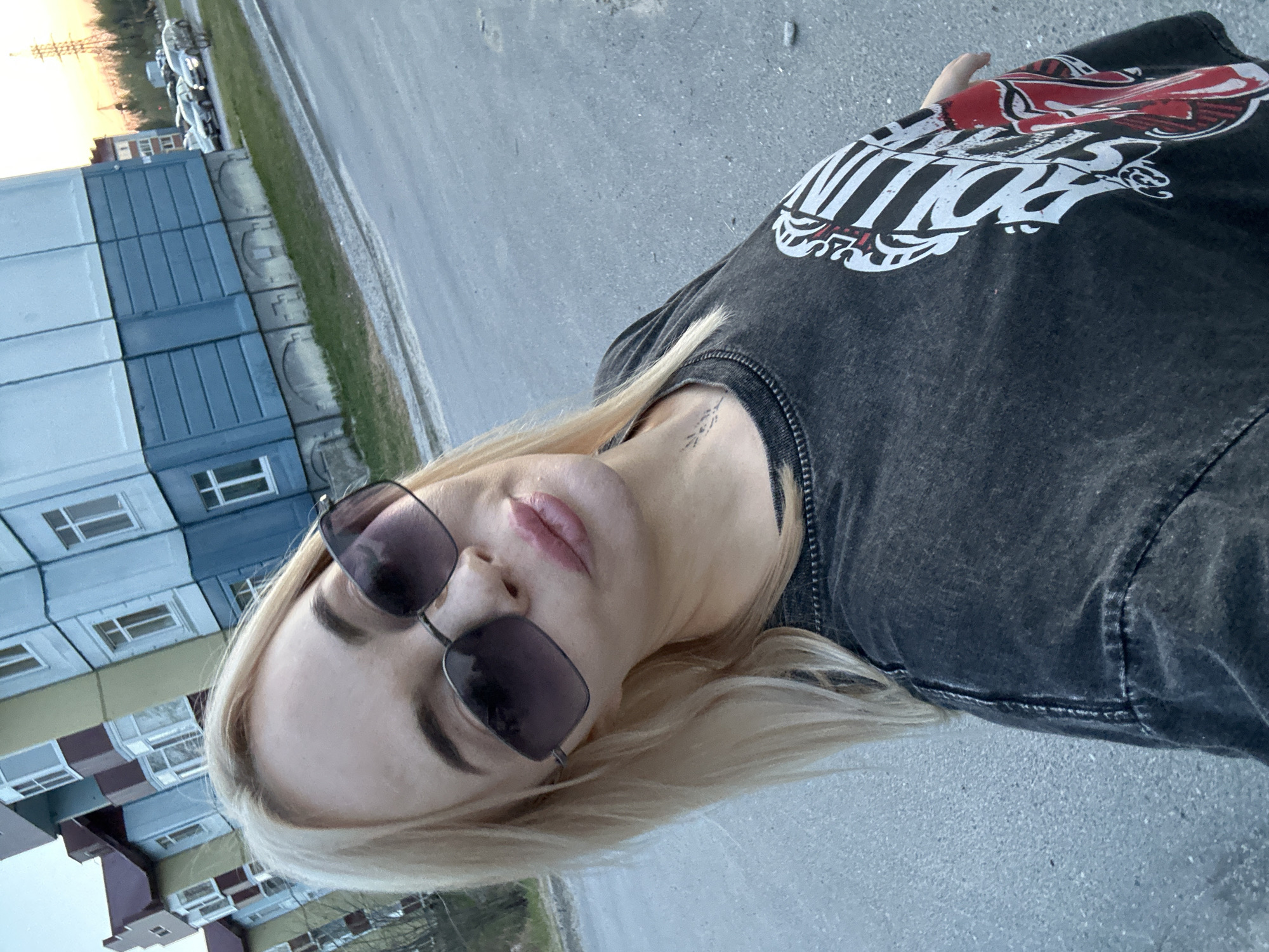 A blonde woman wearing sunglasses and a black t-shirt with a graphic design is the primary subject of this tilted, close-up outdoor shot. Her face is prominent, with a focus on her eyes, lips, and fair complexion. A delicate tattoo is visible on her neck.
The setting appears to be urban. A multistory building constructed of modular sections is in the background. A road runs alongside the building. Beyond, are parked cars. The image suggests the location might be Высокий, Russia. The focus is on the subject's pose and appearance.