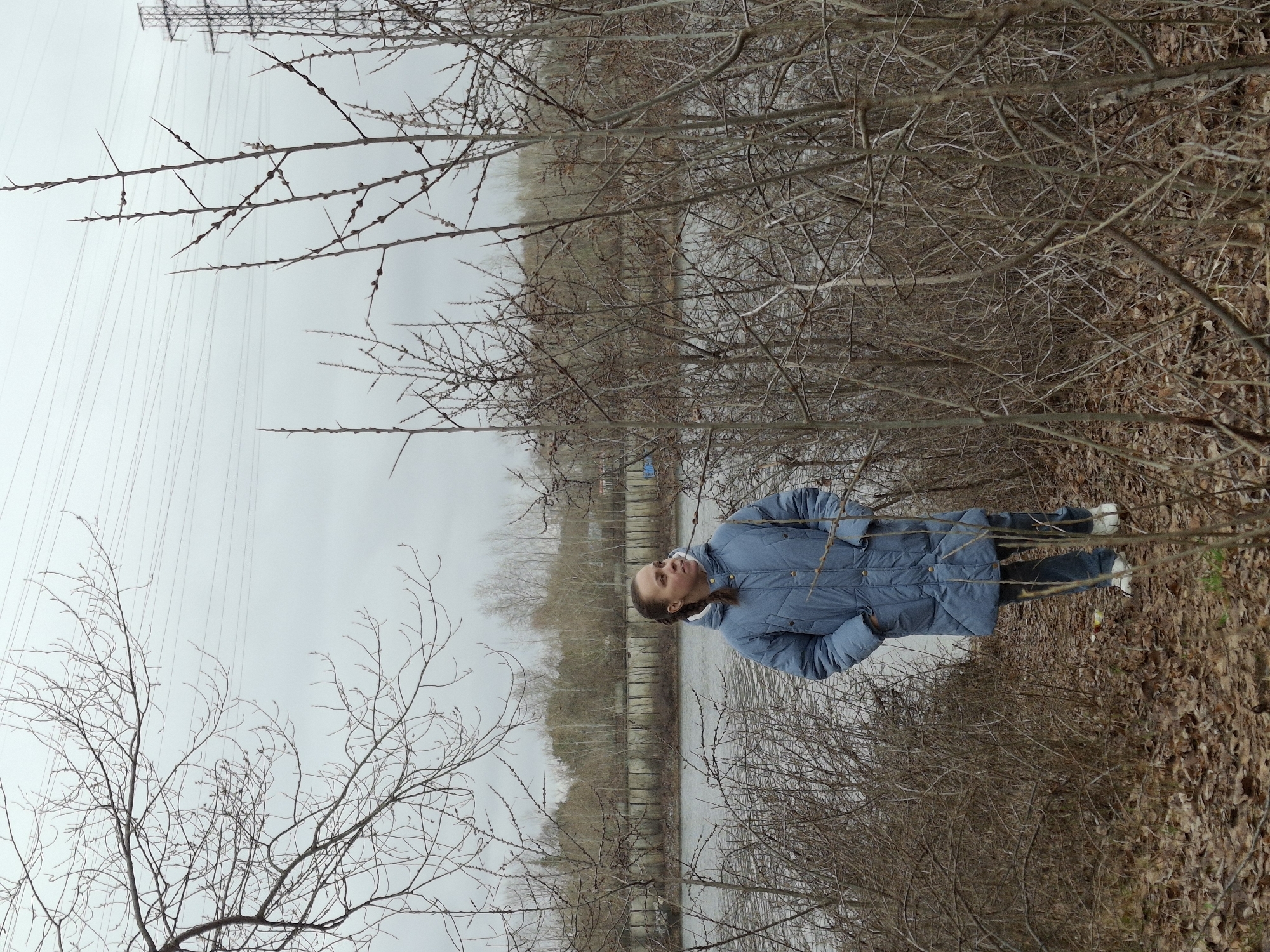 A girl with brown hair in a braid is seen in an outdoor setting near a body of water. She is wearing a blue puffer jacket, dark pants, and white shoes. The landscape includes bare trees, dry leaves on the ground, and a concrete wall along the water's edge. Overhead power lines are visible in the distance. The location is in Chaikovsky, Russia.