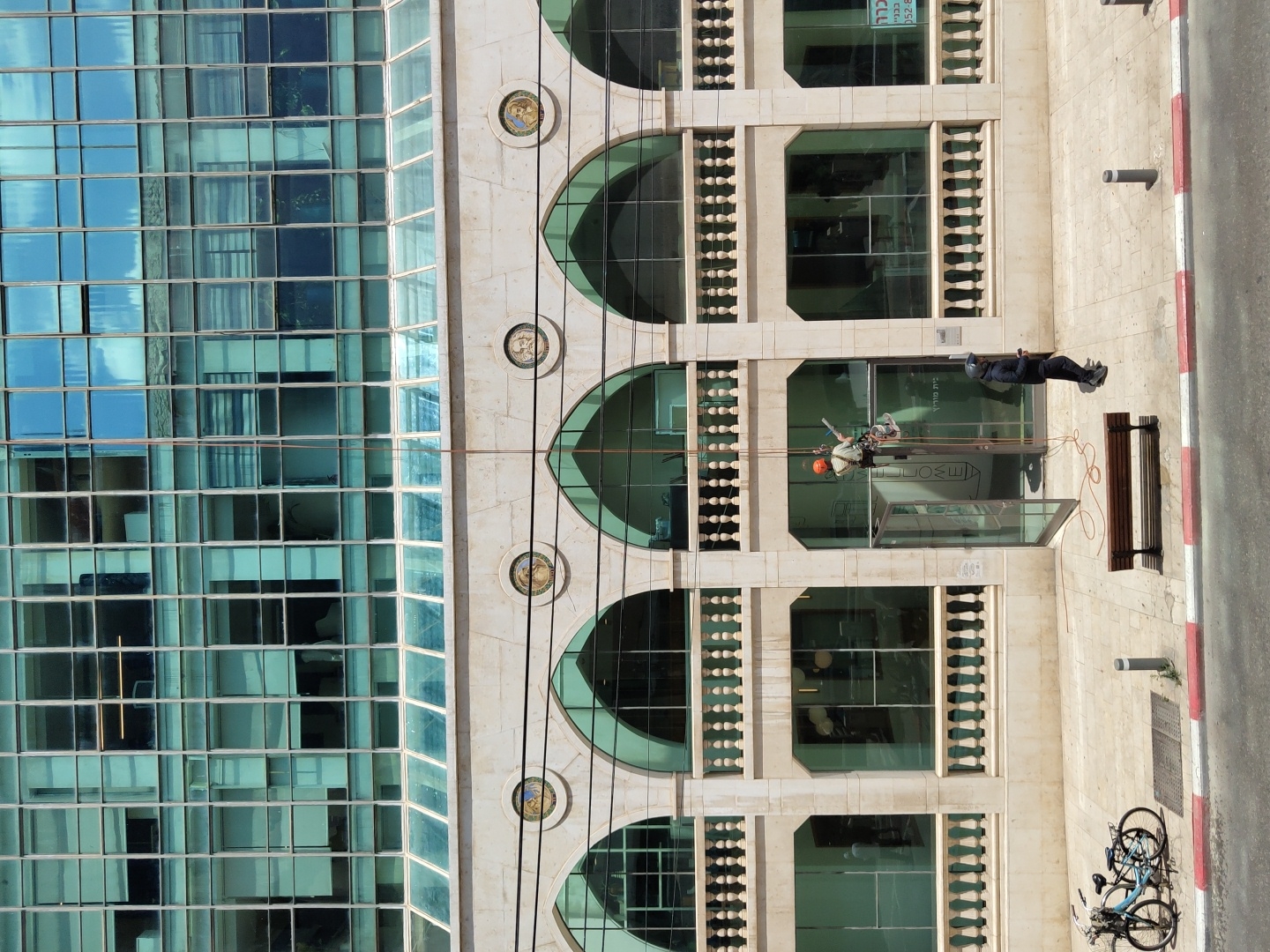 A high-angle, outdoor shot depicts the exterior of a building in Tel-Aviv, Israel, specifically featuring adjacent contemporary and classical architectural styles. The left side showcases a modern glass-curtain-wall building with a grid of windows reflecting the sky. To the right is a classically designed building with a pale stone facade, arched windows, and decorative balustrades. Between the buildings are circular medallions.
Two window washers are suspended on ropes outside a window on the classical building. One worker is wearing an orange helmet, and the other is wearing a white helmet. On the sidewalk below, a worker watches the window washers.
The sidewalk is partially visible, featuring a red and white curb, a wooden bench, two bicycles parked near a metal grate, and a short metal pipe. The street is partially visible.
