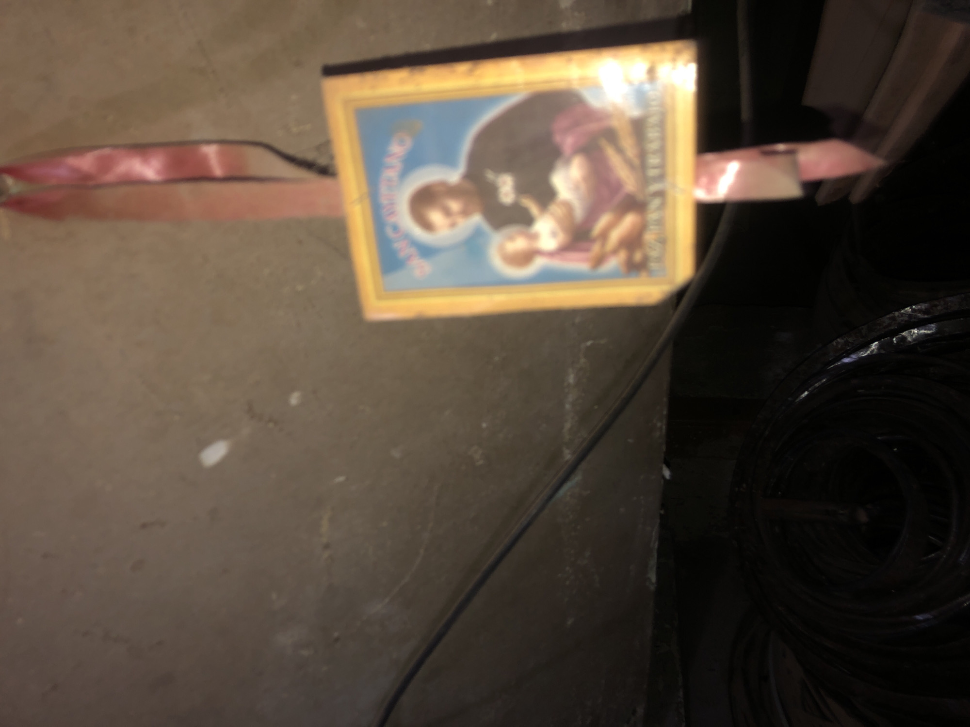 The image shows a religious plaque featuring "San Cayetano," secured with pink ribbon or tape, lying on a dusty, concrete floor. A tightly coiled black hose is visible in the background, partially obscured by shadow. The floor surface is gray and uneven, and a dark, undefined space is present in the upper right corner. The location, Buenos Aires, Argentina, cannot be directly determined from the visual information provided.