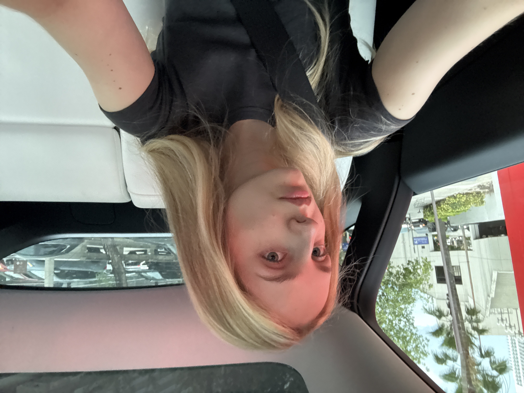 A young Caucasian female with blonde hair and blue eyes is seated in the back seat of a car, looking at the camera. She is wearing a dark gray t-shirt and a black seatbelt. The car's interior is visible, with white leather seats and a sunroof. Outside the car, a cityscape with buildings and palm trees can be seen through the window, suggesting a location in Miami, Florida, United States of America. The lighting suggests it is daytime.