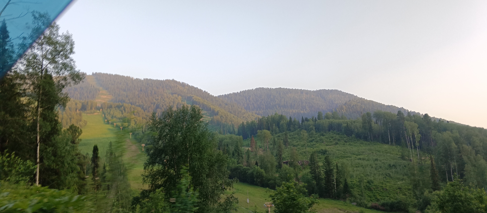 This image depicts a scenic view of a mountainous, forested landscape, likely in Iogach, Russia. The foreground is dominated by lush green trees, with a blurred blue object, possibly a tinted window or filter, occupying the upper left corner.
The main subject of the image is the expansive mountain range covered in dense coniferous and deciduous forests. A ski slope or trail is visible cutting through the trees on the left side of the mountains, with ski lift poles sparsely dotted along its path. The lighting suggests it is either early morning or late afternoon, with a soft, diffused light casting a warm glow on the slopes and highlighting the greenery. The sky is a pale, hazy gradient, indicating potentially overcast conditions or the soft light of twilight.
The overall mood is serene and tranquil, emphasizing the natural beauty of the mountainous region. The untouched wilderness, combined with the man-made ski slope, suggests a recreational area within a natural setting. There is no visible text in the image.