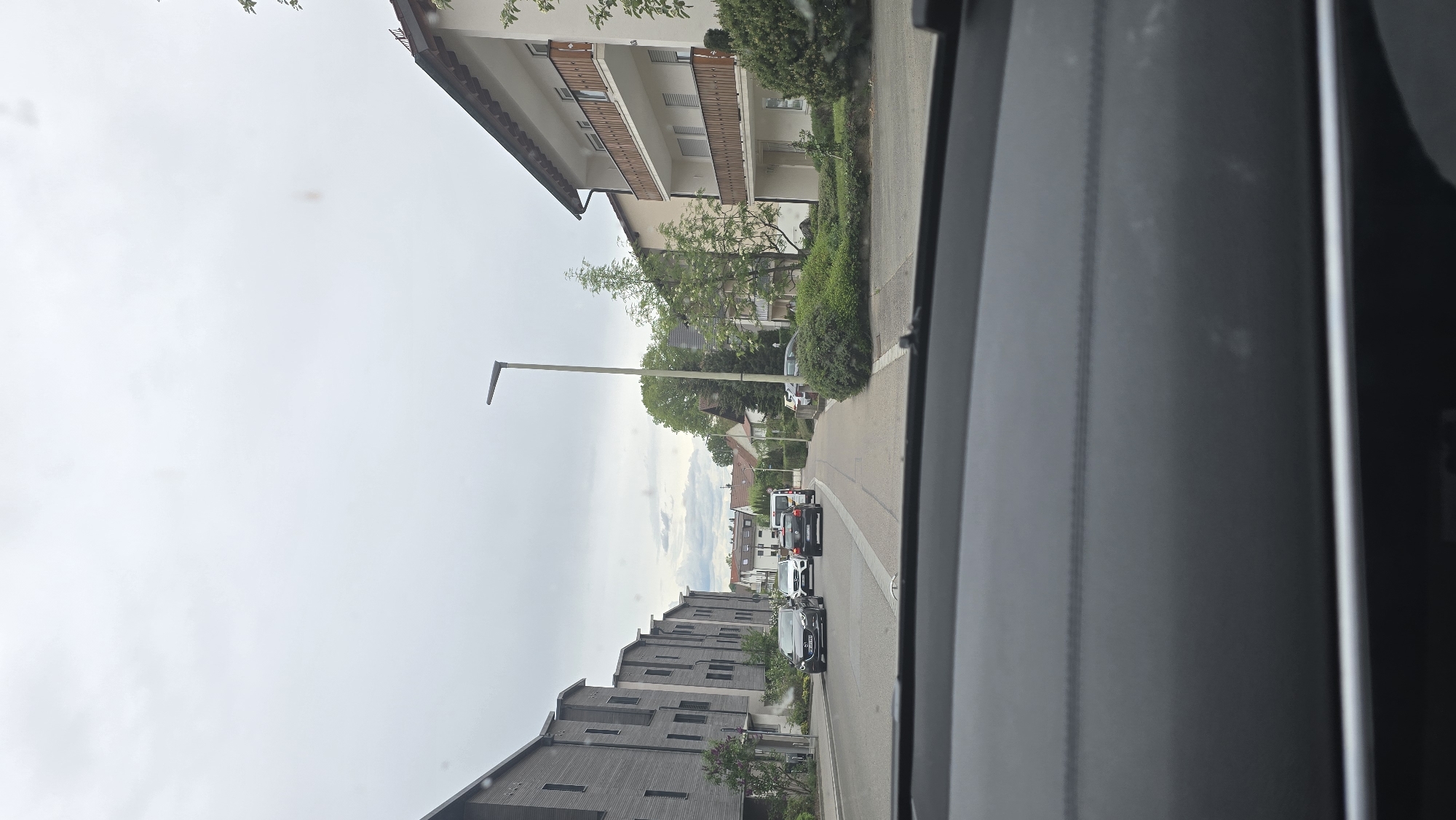 The photograph shows a view from inside a vehicle driving on a street in what appears to be a residential area. The buildings on the right side of the street appear to be apartment buildings with balconies, while the buildings on the left side are modern, grey apartment buildings. Several cars are visible on the road ahead. A streetlight is seen on the left side of the street. The weather appears overcast. The location is Pfaffenhofen an der Ilm, Germany.