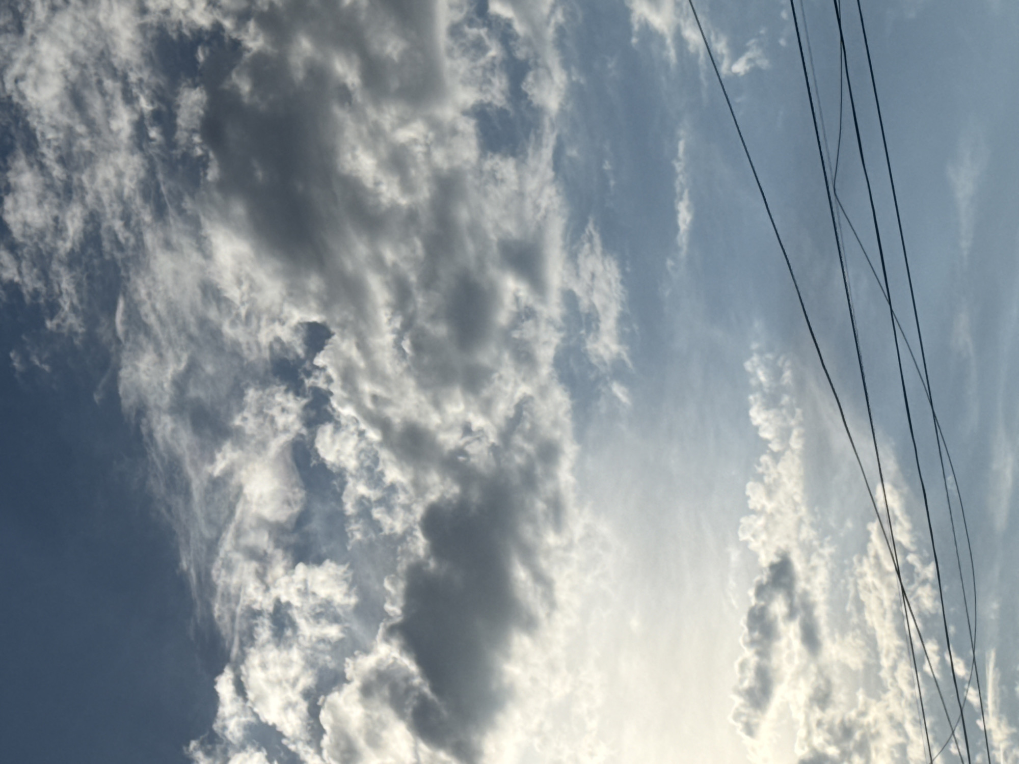 This is an outdoor, vertical, medium-distance shot of a sky filled with clouds and power lines. The sky is a light blue, with white and gray clouds scattered across it. Some clouds are fluffy and bright, while others are darker and more textured. Several dark power lines are visible in the lower portion of the image, crisscrossing each other. The lighting suggests it is daytime, possibly late afternoon or early morning due to the soft light filtering through the clouds. No people or buildings are visible. The image was taken in Abovyan, Armenia.