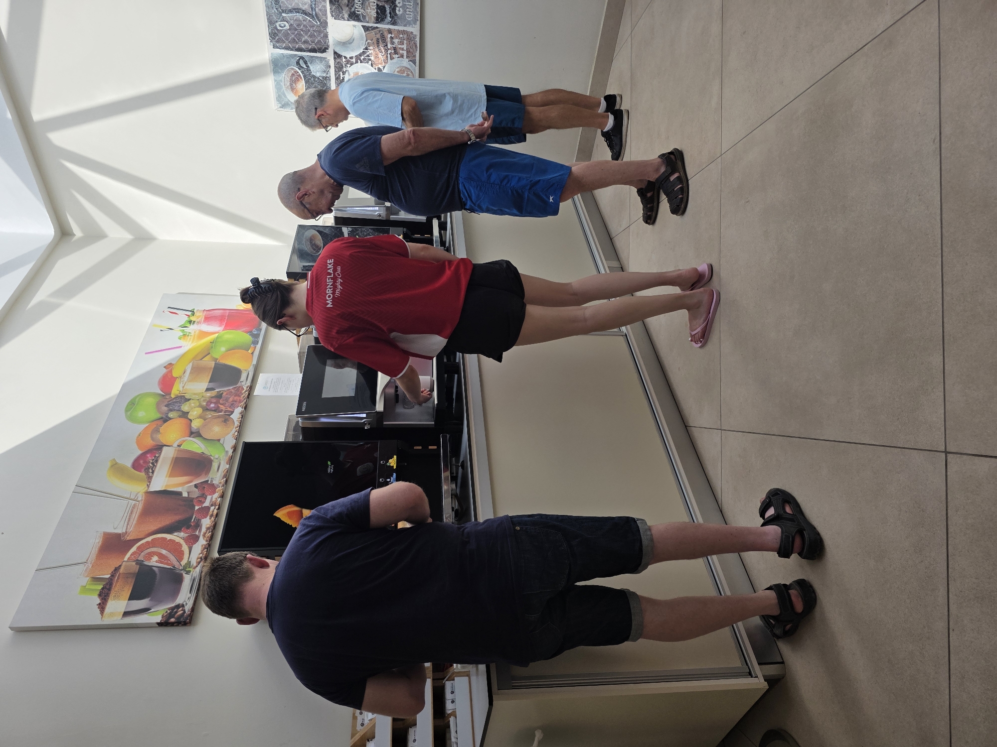 In Costa Teguise, Spain, four individuals are standing in a line at a self-service drink station in what appears to be a hotel or resort. The person at the front, a man wearing a dark blue t-shirt and denim shorts, is facing away from the camera and interacting with a drink dispenser. To his right, a woman in a red shirt with the "MORNINGLAKE Mighty Oats" logo and black shorts is also at the dispenser. Next in line is a man in a dark blue t-shirt and blue shorts, looking down. The last person in line, a man in a light blue t-shirt and blue shorts, is also looking at the dispensers. The drink station features multiple chrome and black machines, one of which has a screen displaying an orange slice. Above the station, a large panoramic mural depicts various fruit juices and coffee drinks. The floor is tiled in a beige, square pattern, and the environment is well-lit with natural light streaming from above, casting shadows on the wall.