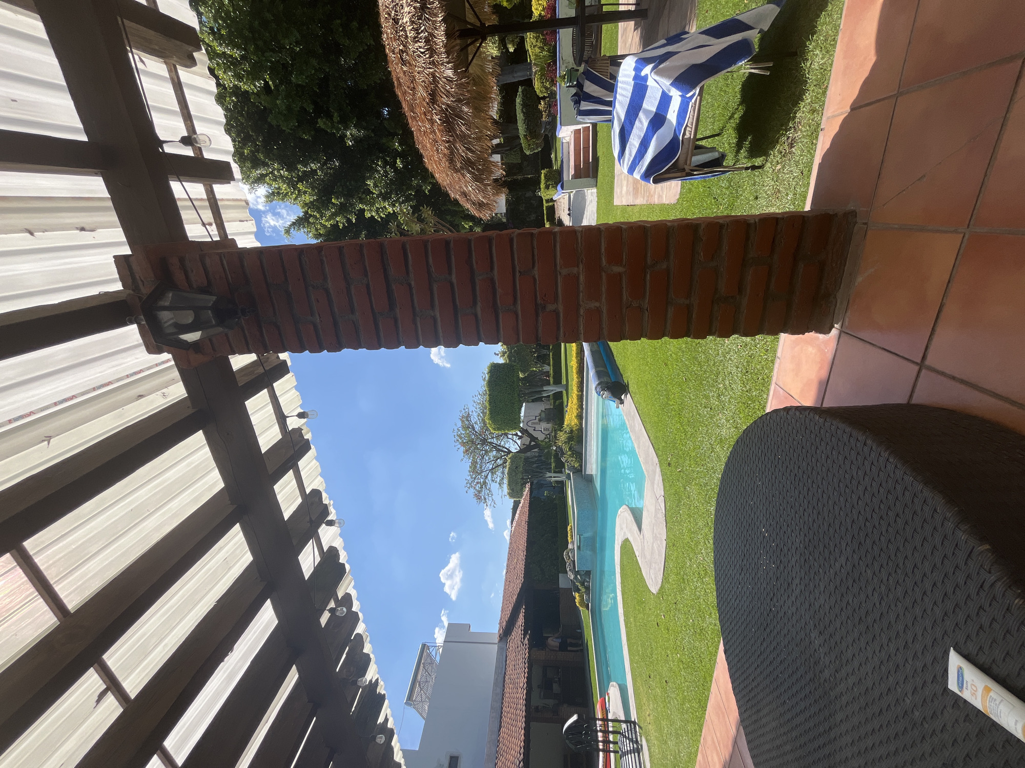 The image shows an outdoor residential scene with a swimming pool and patio area in Unknown, Unknown. The foreground contains a brick pillar supporting a translucent roof structure with string lights. A dark wicker table is partially visible on the right. Beyond the pillar, a lawn extends to a curved swimming pool. A white building is visible in the background, and a thatched-roof umbrella is partially seen to the left. A blue-and-white striped towel is draped over a chair near the thatched-roof umbrella. The sky is visible above the buildings, with a few scattered clouds.