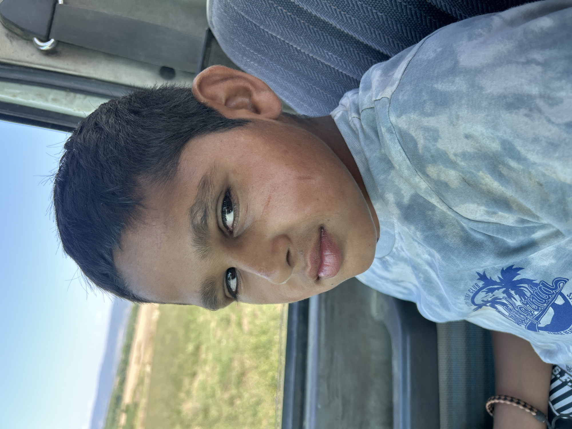 A young boy with dark hair and brown eyes is seated in a vehicle. He is wearing a light blue tie-dye t-shirt with a blue graphic design on the front. The boy is looking towards the camera with a neutral expression. Outside the vehicle, a grassy field and a distant hill are visible under a clear blue sky. The interior of the vehicle shows a grey fabric seat and part of the door panel. The image is taken from a close-up perspective, focusing on the boy's face and upper body. There is no visible indication of the location being Poroy, Bulgaria.