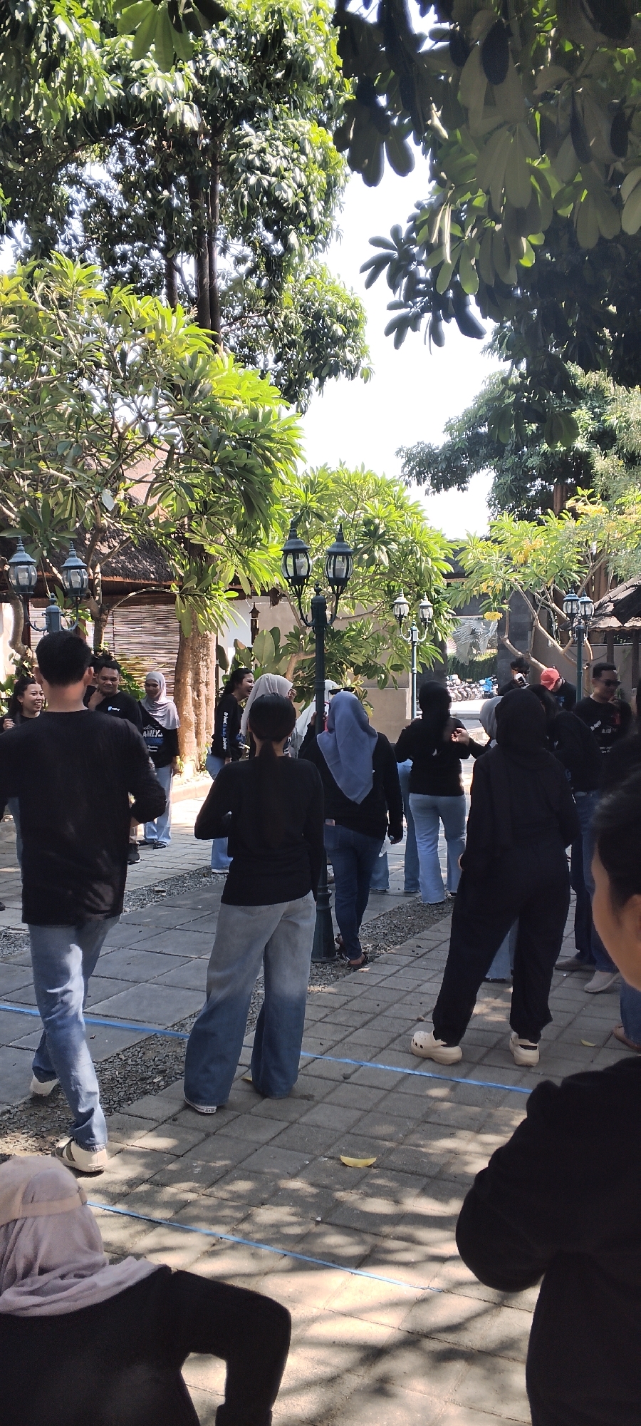 The image captures a group of people gathered outdoors in what appears to be a communal area with lush trees and paved walkways. The setting suggests a warm, possibly tropical climate, with sunlight filtering through the foliage. The people are dressed in casual attire, with several individuals wearing black shirts or jackets and jeans. Some are wearing headscarves, indicating a possible cultural context.

The arrangement of the individuals suggests they might be participating in an activity or event. Some are facing away from the camera, looking towards a central point, while others are engaged in conversation. The environment features decorative lamps on poles, adding a touch of aesthetic to the outdoor space. The ground is a mosaic of gray paving stones interspersed with gravel.

The mention of Talise, Indonesia, places the scene in a specific geographical location, hinting at the architectural style of the buildings in the background, which have thatched roofs, a common feature in Indonesian vernacular architecture. The presence of parked motorcycles in the background further reinforces the Indonesian context. The overall impression is one of a casual social gathering or team-building activity taking place in a well-maintained outdoor space.