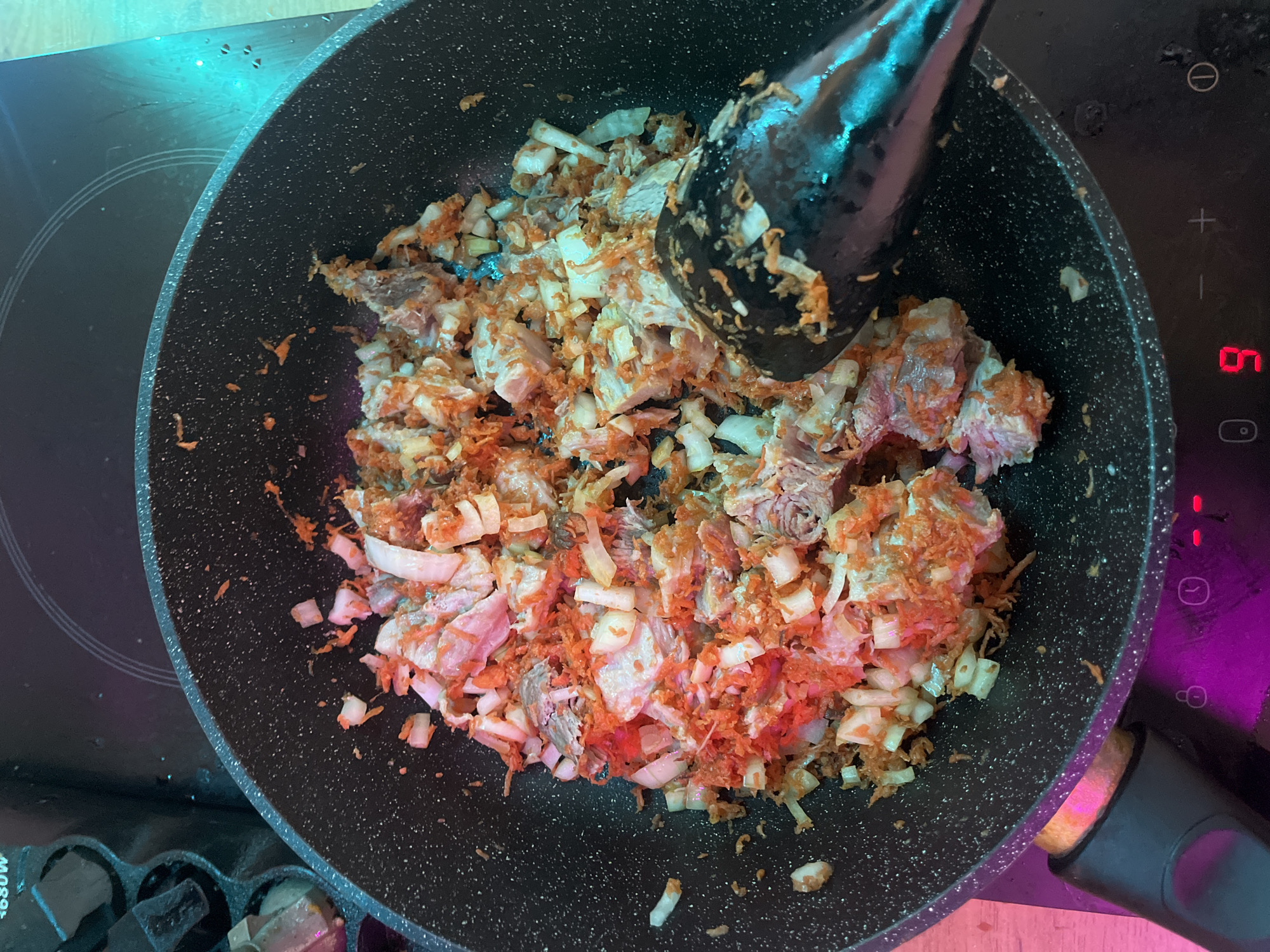 A close-up, overhead shot shows ingredients being cooked in a black non-stick frying pan on a modern black induction hob. The ingredients appear to be diced pork, shredded carrots, and chopped onions, mixed together and starting to brown. A black spatula is partially visible in the lower right quadrant of the pan, stirring the food. The induction hob has a digital display showing a "9" and other control icons. The background is dark, with some hints of colored lighting, likely from nearby electronics or ambient light sources. There is no indication of the specific location within Saint Petersburg, Russia.