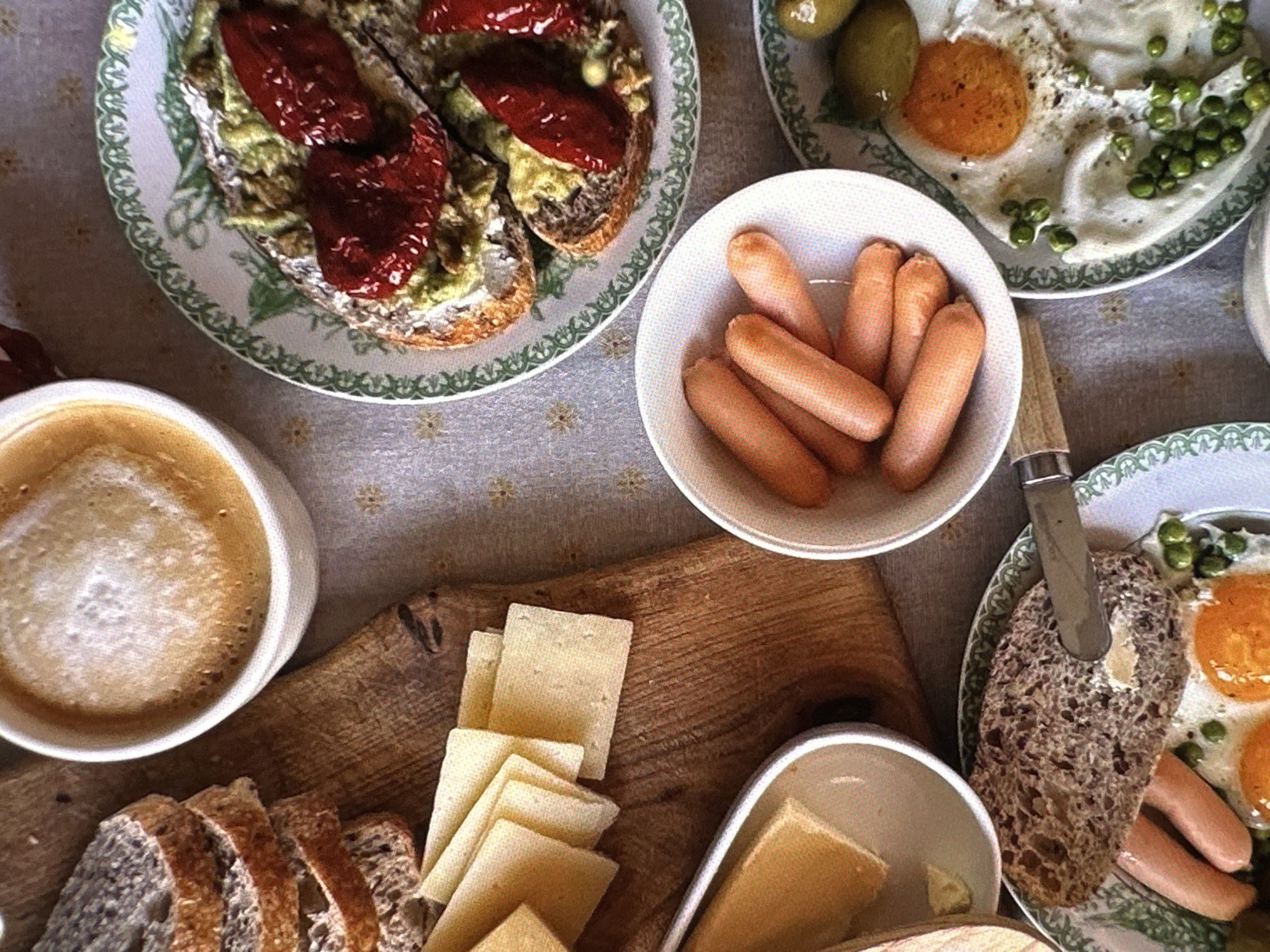 The image shows a tabletop laden with food, presenting a meal setting. There are plates with various items including what appears to be avocado toast with sundried tomatoes, fried eggs with peas and olives, and a bowl filled with small sausages. A wooden board holds sliced bread and cheese. A cup of coffee with froth sits to the side. The location is not determinable from the image. There are no people visible in the image. Actions and interactions are not observable, but the scene suggests someone is preparing to, or has just, eaten. The environment appears to be indoors based on the lighting and arrangement.