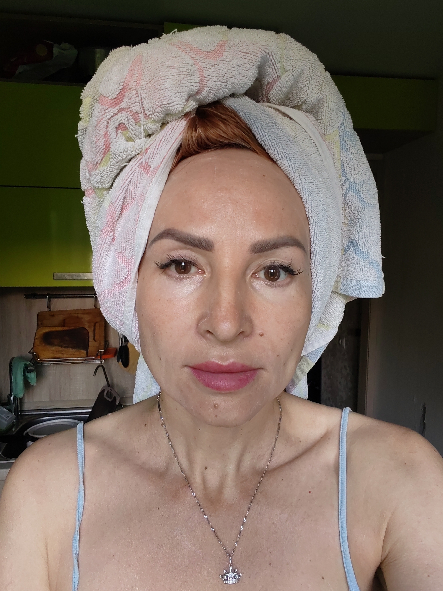 A medium shot shows a woman with auburn hair wrapped in a white towel with pink and blue stripes. She is looking directly at the camera. Her skin is fair, with some freckles and a few blemishes. Her eyes are brown, with long, dark eyelashes. Her lips are a light pink, and she is wearing a silver necklace with a crown-shaped pendant. She is wearing a light blue camisole. The background appears to be a kitchen. A green cabinet is visible on the left, with a wooden cutting board hanging on the wall. The image was taken in Chaikovsky, Russia.