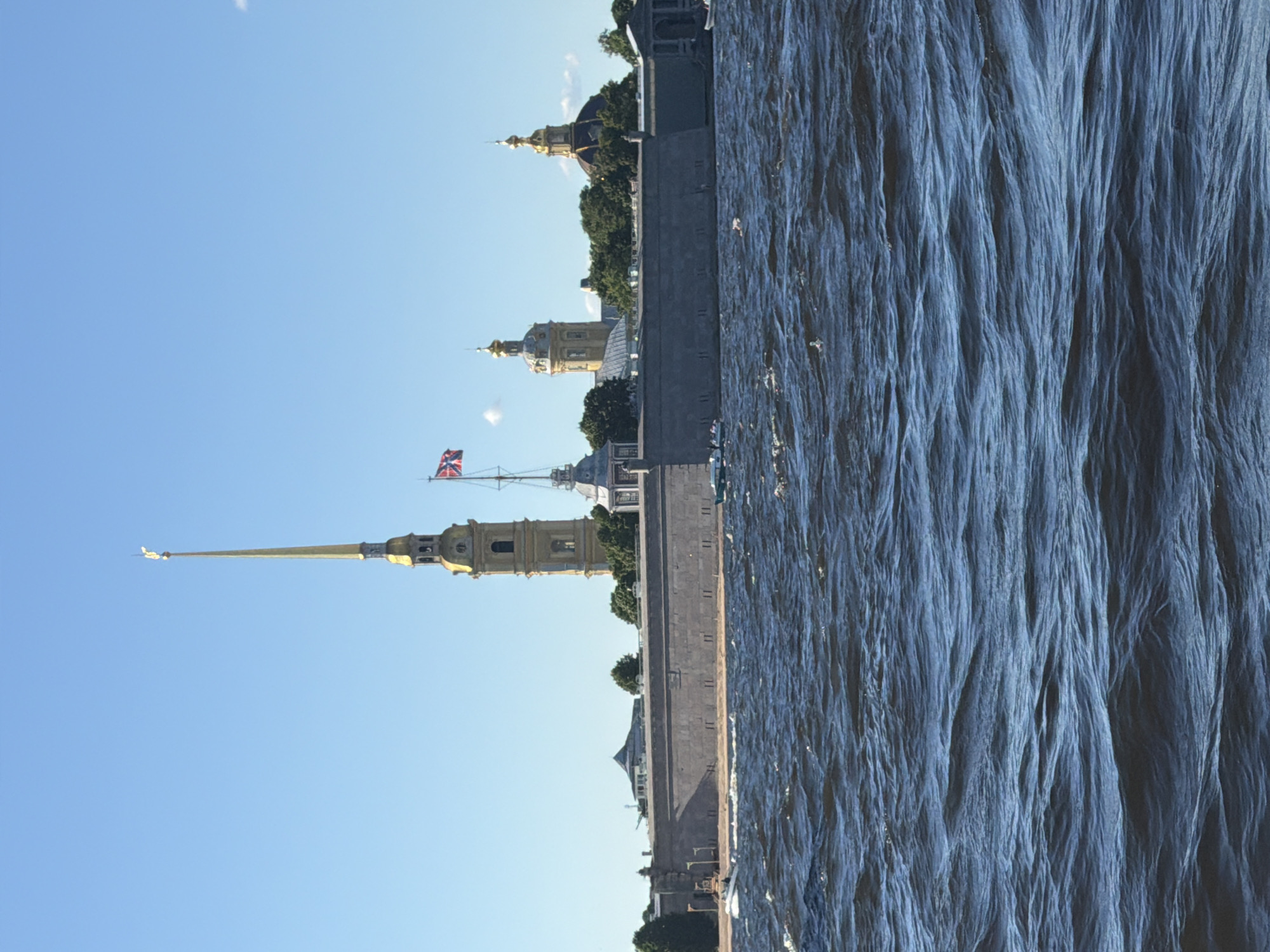 This is a medium shot, eye-level, outdoor, daylight, vertical, photograph of the Peter and Paul Fortress in Saint Petersburg, Russia. The fortress wall is in the middle ground, running horizontally across the frame. A tall, slender, golden-domed spire with a cross at the very top rises from behind the wall on the left side of the image. To the right of the spire, a Russian flag flies on a mast. Several other golden-domed buildings are visible behind the fortress wall. The water in the foreground is dark blue and choppy, with small whitecaps. Two small boats are visible in the water near the fortress wall, one white and one green. The sky is clear and blue.
