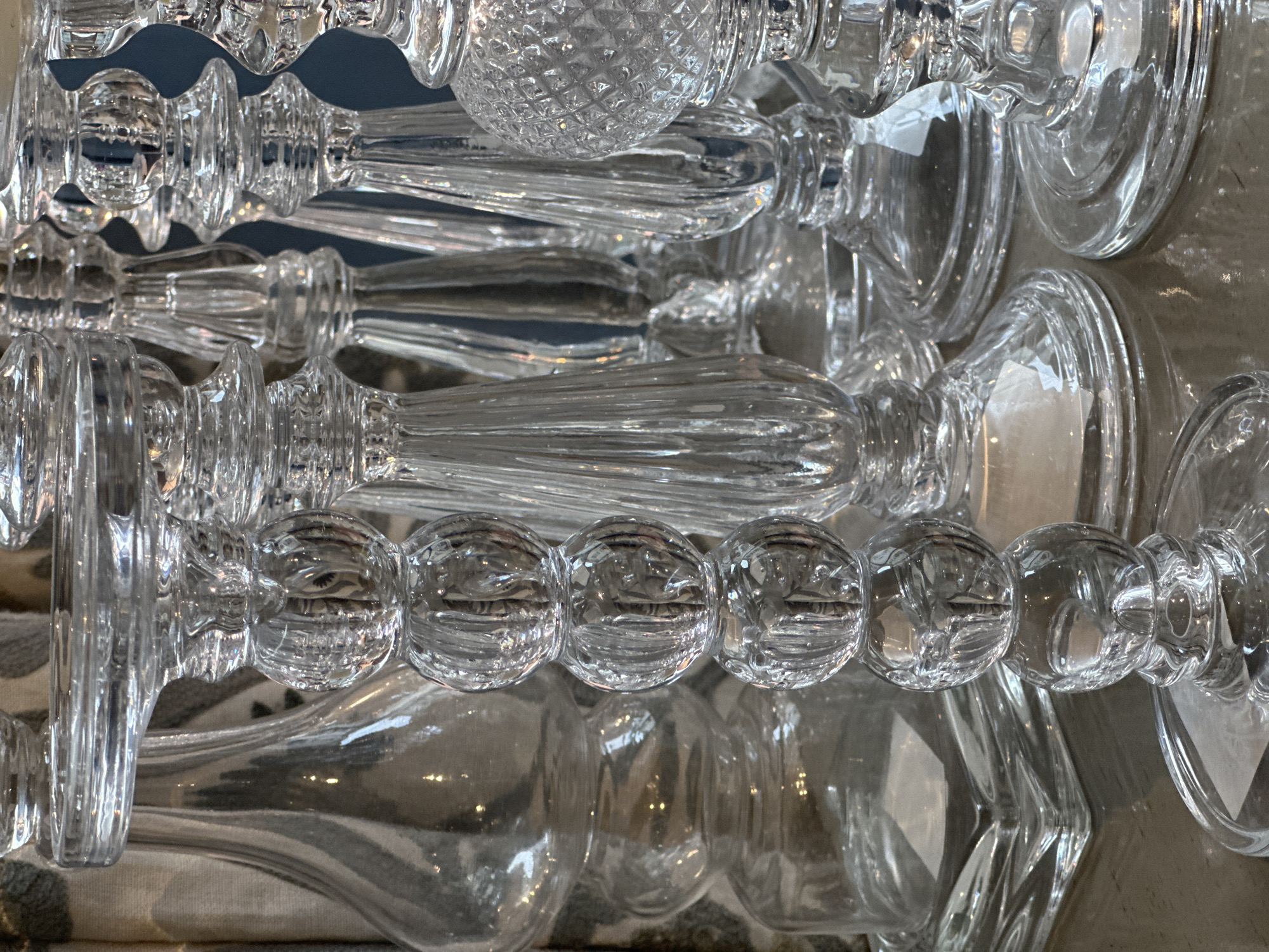 A close-up shot features multiple transparent glass objects with intricate designs, likely the stems of glasses or decorative items. The objects are arranged overlapping each other, creating a complex pattern of reflections and refractions. Some items have spherical or bulbous elements, while others are more elongated and textured. The background is slightly blurred and out of focus, but the overall composition is focused on the details and textures of the glass objects. It is challenging to determine the exact location within Tunbridge Wells without additional information or context.