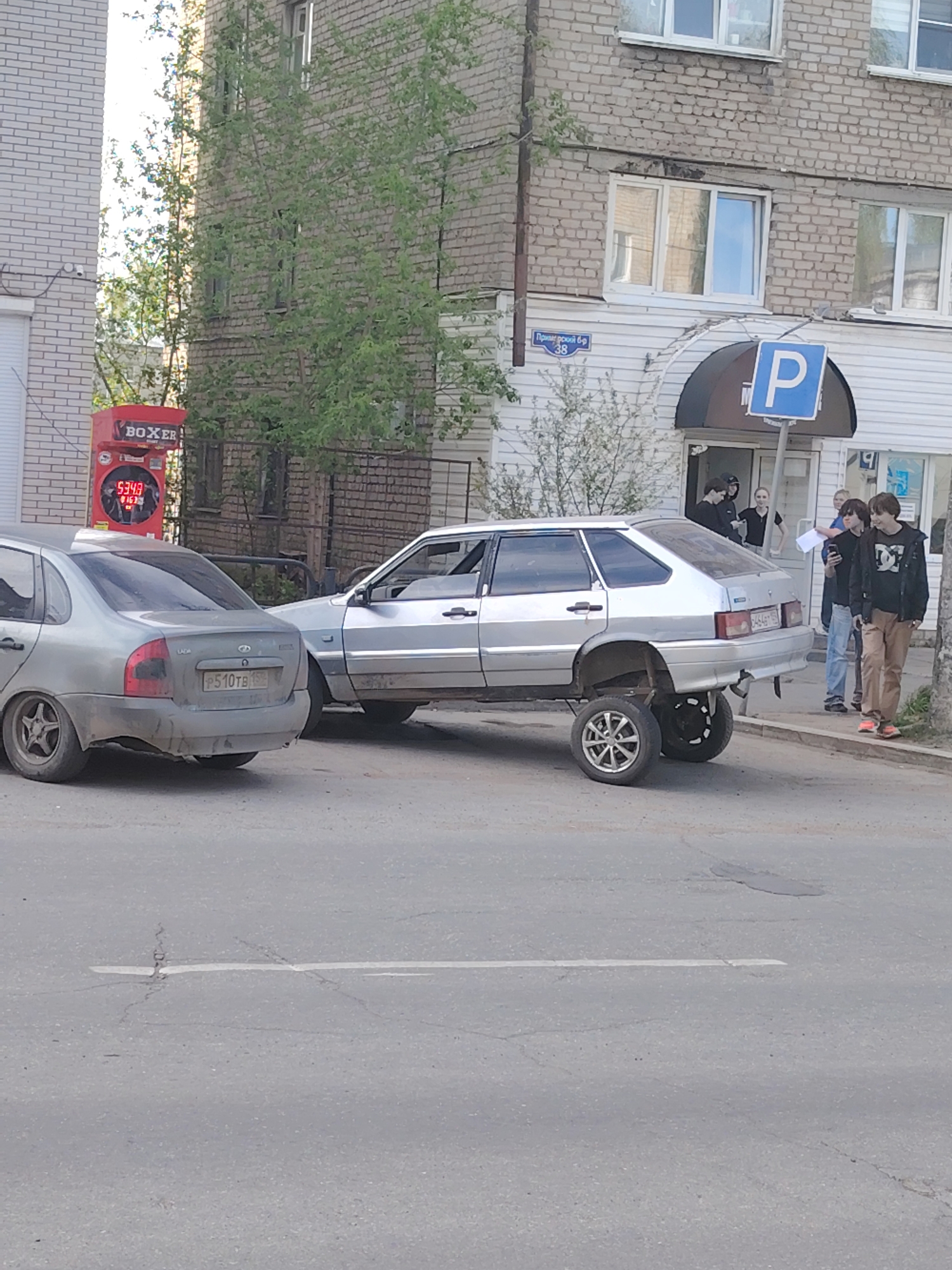 The image shows two cars on a street in what appears to be a residential or commercial area of Chaikovsky, Russia. The car on the left is gray and appears to be a LADA. The license plate on this car reads P510TB 15. To the right, a light-colored car sits elevated on what looks like two stacked tires each, with a total of four tires supporting the rear of the vehicle. Several people are standing near the car that's been lifted, possibly observing or discussing the situation. A blue "P" parking sign is visible in the background near a building labeled "Примерский б-р 38". A Boxer electronic boxing game kiosk sits next to the gray car on the left. The surrounding environment includes buildings, trees, and a street.