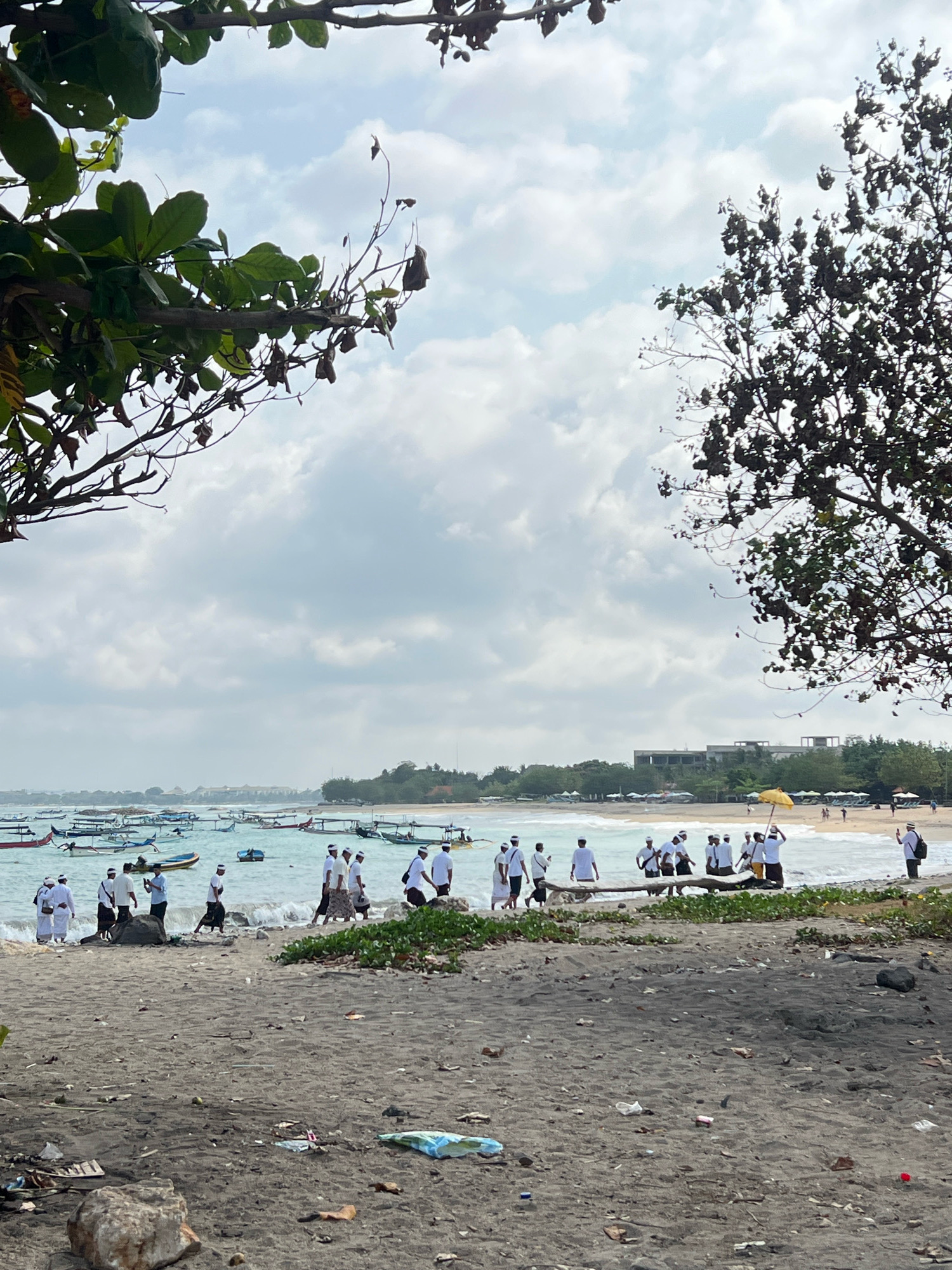 The image shows a beach scene in Kuta, Indonesia. In the foreground, a sandy beach is littered with various pieces of trash. Further back, a group of people dressed in white shirts and dark skirts, many with white headbands, are walking by the water's edge. They appear to be participating in a procession or ceremony. Beyond them, the ocean is filled with numerous traditional Balinese fishing boats. In the background, a coastline with palm trees, resort buildings, and a clear sky with scattered clouds is visible. The overall impression is a lively beach with cultural activity.