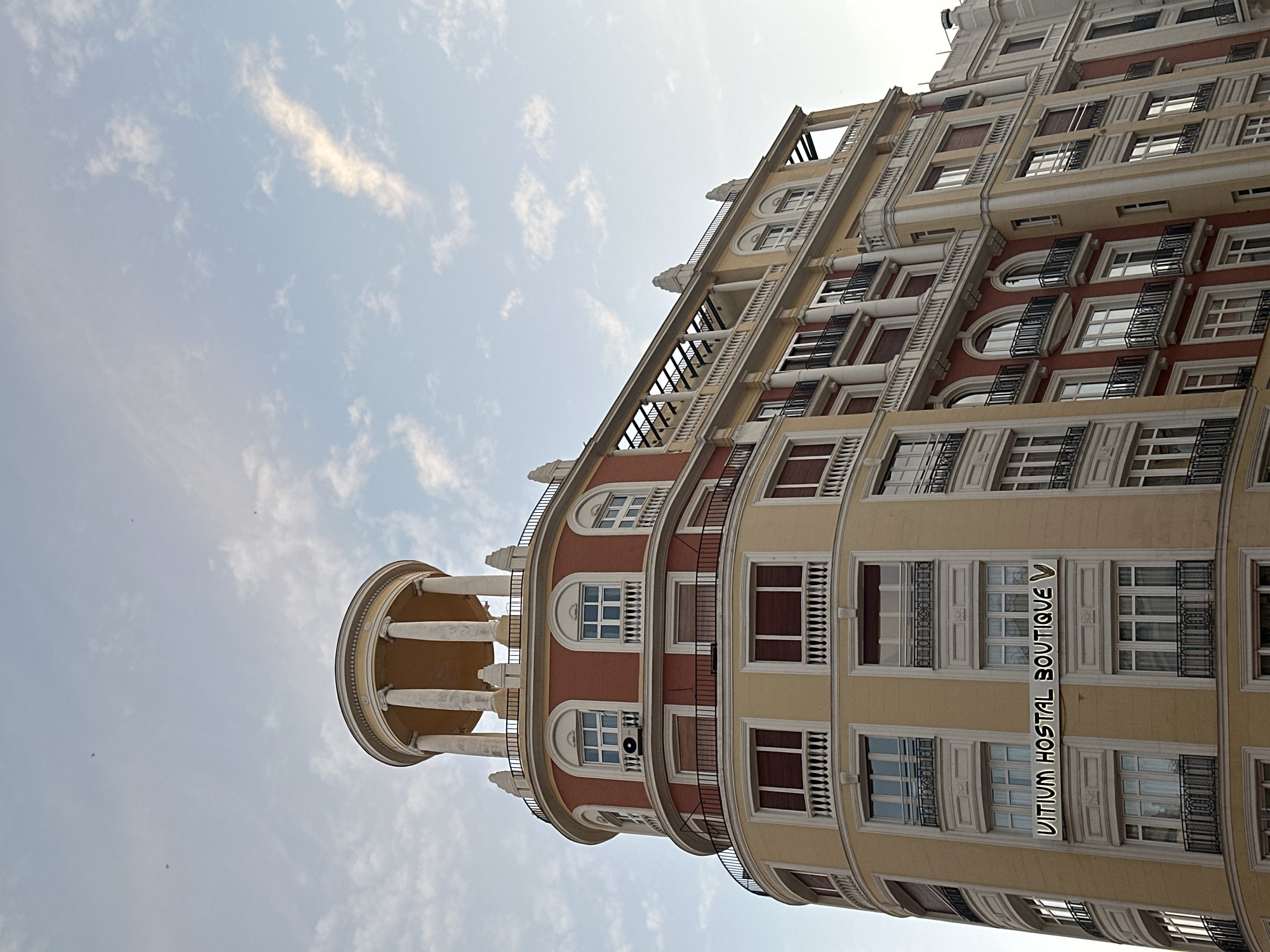 The image shows a portion of a tall building in Madrid, Spain. It features a distinctive cylindrical tower topped with a colonnade, located on the lower left of the frame. The building has multiple floors with windows, balconies, and decorative architectural elements in a palette of pale yellow and reddish-brown. The building appears to have some commercial purpose, as visible on the lower levels the text "VITIUM HOSTAL BOUTIQUE V" is visible. The background shows a pale blue sky with light clouds. The perspective is looking upwards at the building, which is positioned at an angle.