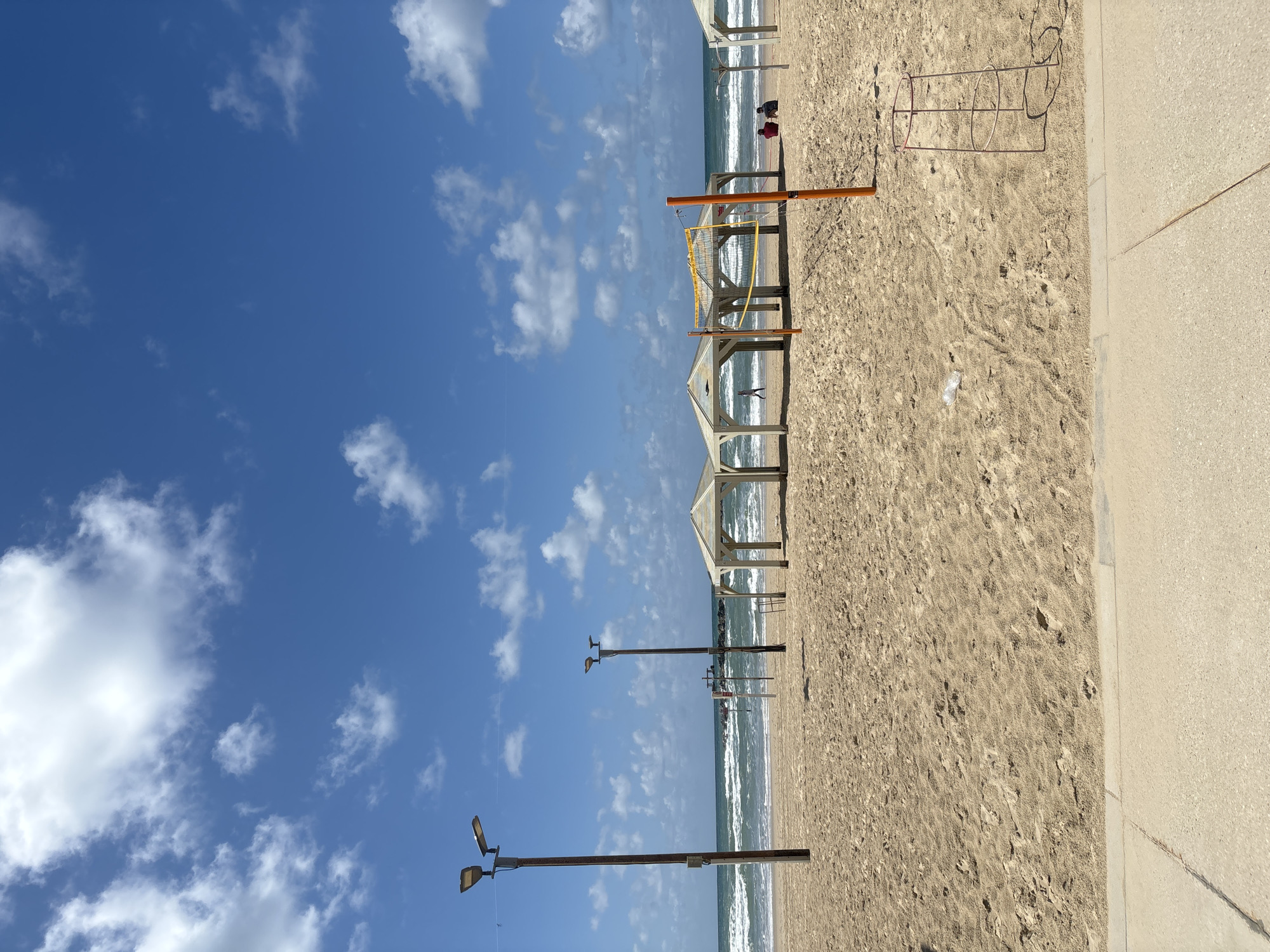 This image shows a beach scene in Tel Aviv, Israel. The beach has light-colored sand and the Mediterranean Sea is visible in the background. Several white beach canopies are arranged in a line along the beach. A volleyball net stands near the canopies. A person or people are sitting on the beach near the shoreline. Light poles are visible in the distance. A sidewalk or paved area runs along the right side of the image. The sky is blue with scattered white clouds. A pink metal structure sits on the sand.
