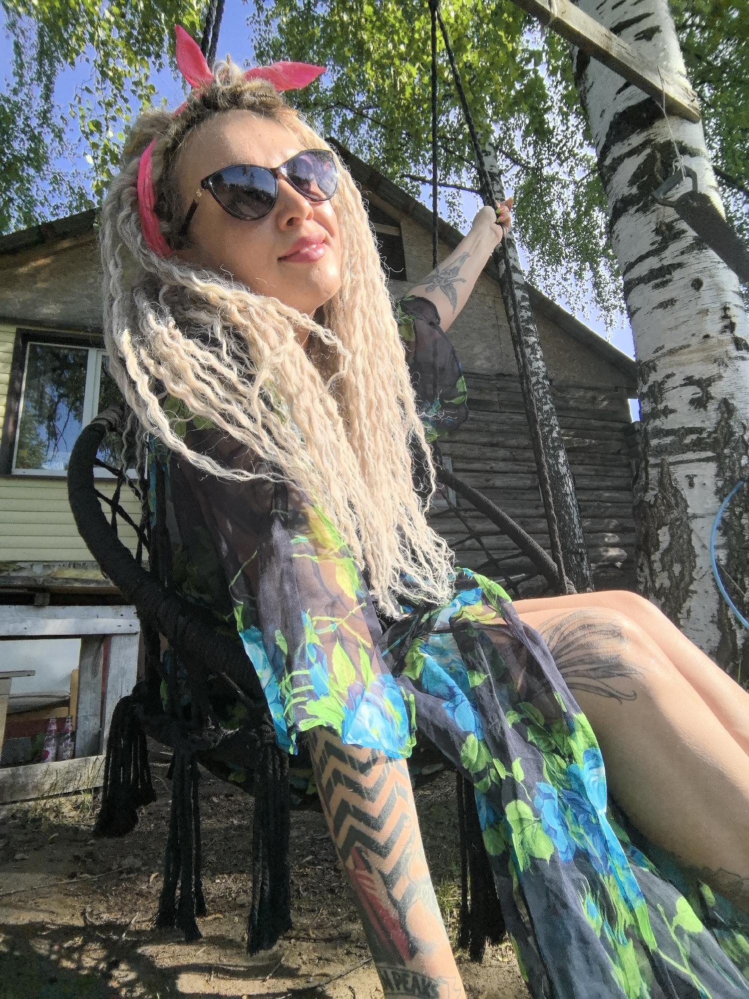 The image features a woman with blonde dreadlocks and sunglasses sitting in a rope swing. She wears a floral print dress and a pink headband with rabbit ears. She has tattoos on her arms and legs. The swing is suspended from a tree, likely a birch, with bark visible. Behind her is a weathered wooden structure, possibly a house or shed. The setting appears to be outdoors, on a sunny day. No specific city is confirmed.