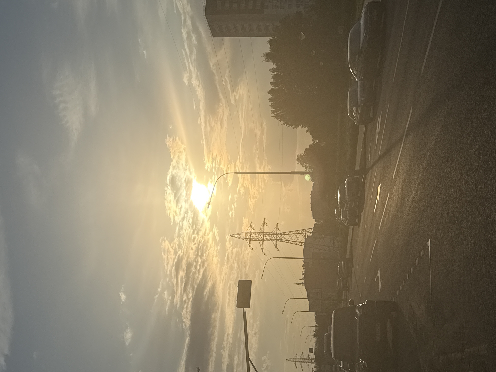 The image shows a street scene in Khimki, Russia, during bright sunlight. The camera appears to be at street level, possibly from a moving vehicle. The focus is on the road, with several cars parked along the side. A pedestrian walkway or sidewalk is visible. There are streetlights lining the side of the road, and an overhead power line structure is visible. In the distance, a multi-story building and trees are seen. The sky is partially covered with clouds and the sun is high in the sky, creating a strong glare.