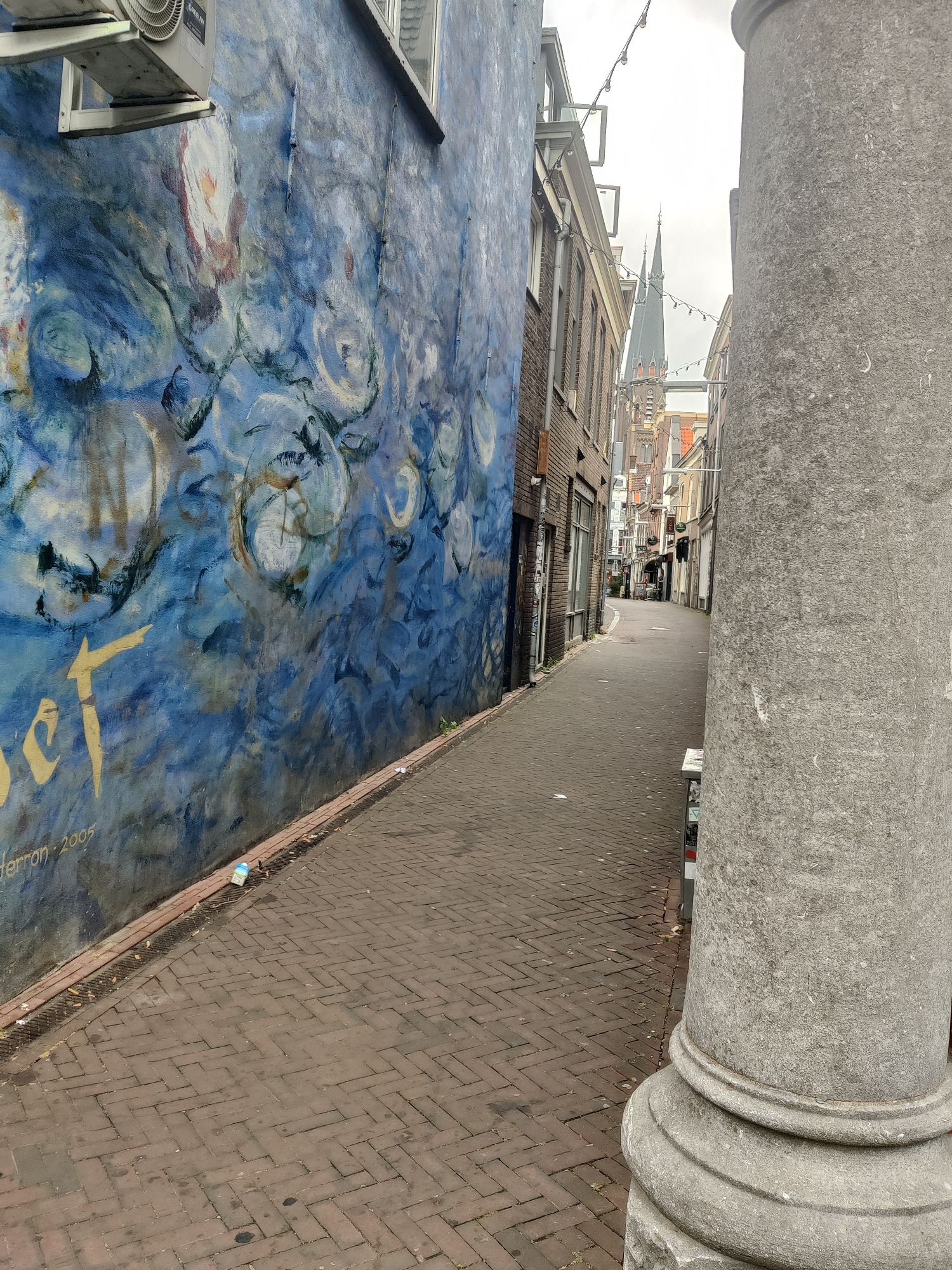 The image depicts a narrow street in Delft, Netherlands. On the left is a building with a large, blue mural featuring abstract circular and floral motifs in shades of blue, white, and hints of pink and yellow. A section of the mural appears to be a stylized yellow inscription. An air conditioning unit is mounted high on the wall above the mural. The street is paved with red-brown brickwork laid in a herringbone pattern. A large, grey, stone column, possibly part of an architectural element, dominates the right side of the frame. In the background, the street recedes between brick buildings, with a tall church spire visible in the distance. Decorative string lights are hung across the street. A single plastic bottle is visible on the ground near the gutter. The overall impression is of an urban alleyway in Delft.