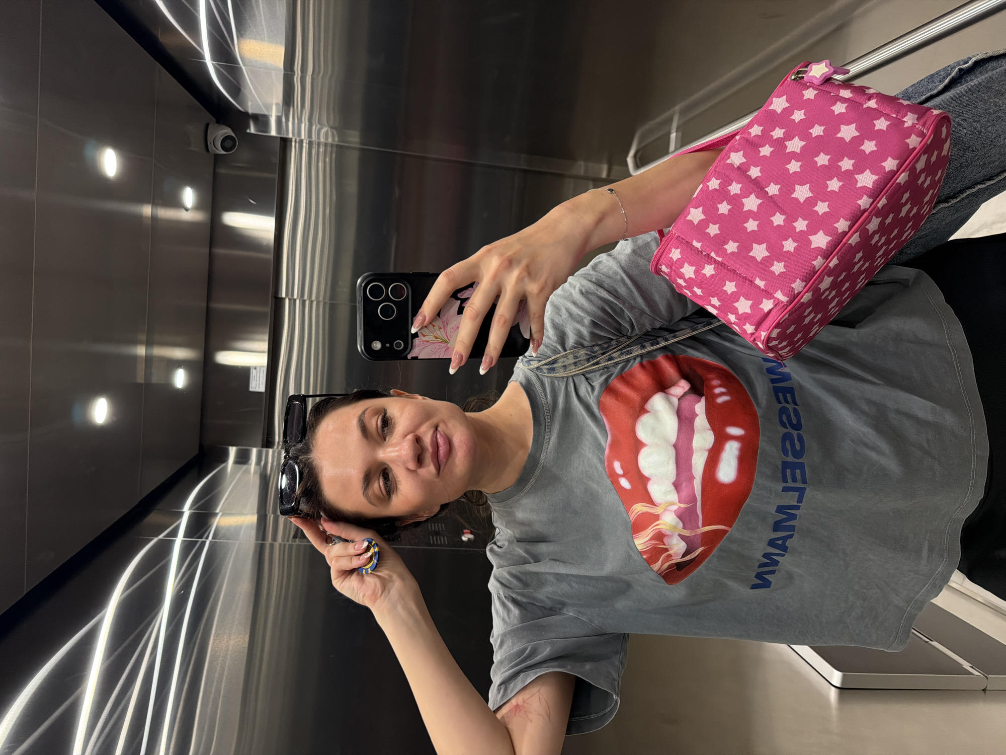 A woman is taking a selfie in an elevator. She is holding a black phone with a floral case in her right hand. Her left hand holds a pair of sunglasses. She is wearing a grey t-shirt with a large graphic of lips on it, a bracelet on her right wrist, and has manicured fingernails. A pink star-patterned bag hangs over her shoulder. The elevator's interior is stainless steel, with lighting fixtures and a security camera visible on the upper portion of the back wall.