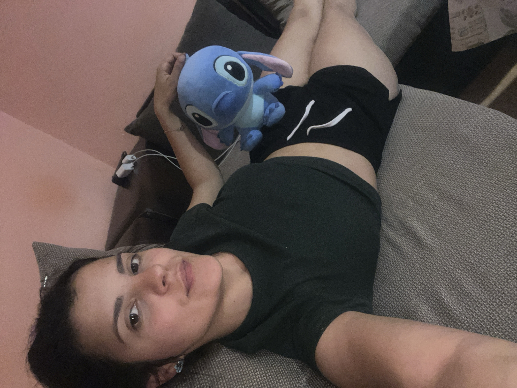 A young woman is depicted lying on a grey couch or bed in an indoor setting, likely in Plovdiv, Bulgaria. She wears a dark t-shirt and black shorts featuring a white stripe detail on the left leg, suggesting she is relaxing or resting. A blue plush toy, resembling the character Stitch, is placed on her torso, and her right hand is gently holding or resting near it. A subtle bracelet adorns her right wrist. In the immediate vicinity of her right arm, white charging cables and a power adapter are visible. The background reveals a light pink wall. The woman has dark hair and eyes, and her expression is neutral to slightly smiling as she looks directly at the camera. The overall lighting is dim, indicating an indoor environment, possibly during the evening or night. No discernible text is present in the image.