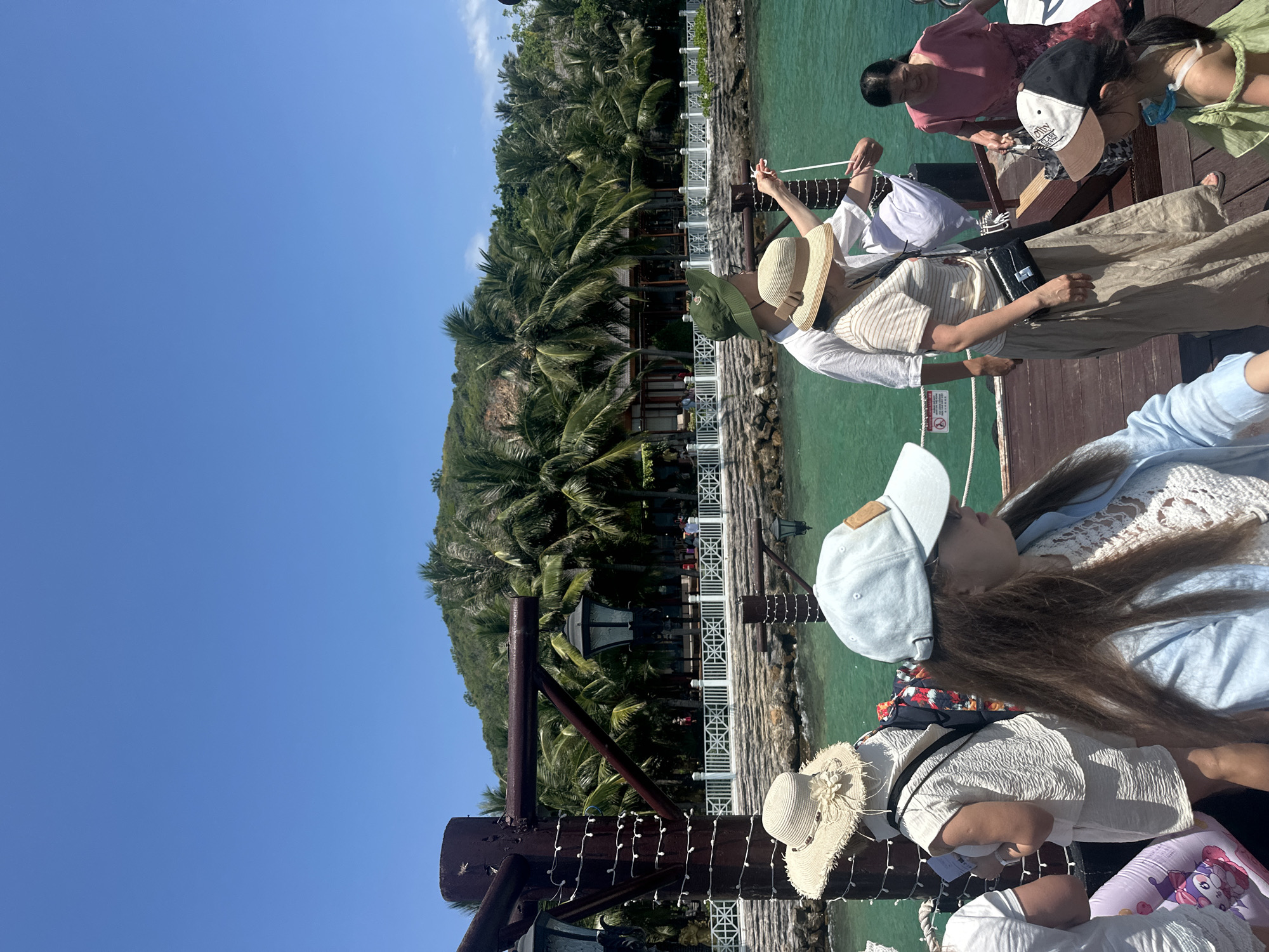 A forensic examination of the image reveals a bustling scene on a wooden pier or boat deck in Nha Trang Ward, Vietnam, under a clear, bright blue sky. The environment is characterized by calm, turquoise water bordering a lush green hillside densely covered with palm trees. In the mid-ground, a white balustrade fence separates the water from a stone retaining wall, behind which a complex of buildings with brown roofs and white facades is visible, integrated into the tropical foliage.
The pier features several dark brown wooden posts, some wrapped with white string lights and others adorned with dark, ornate lanterns.
Approximately ten individuals are present on the pier/deck, mostly adults and at least one child, all dressed in casual summer attire. From left to right in the foreground:
- A partially visible person in a white top, next to a purple and white inflatable ring.
- A person viewed from behind, wearing a wide-brimmed straw hat with a floral embellishment, a white textured short-sleeved top, and matching shorts, carrying a dark shoulder bag.
- A woman with long brown hair, wearing a light blue baseball cap, dark sunglasses, and a light blue long-sleeved top over a white lace garment. She is looking towards the right side of the frame.
In the mid-ground:
- A man wearing a dark green bucket hat and a white t-shirt, standing and holding a white rope, suggesting an interaction with a vessel or mooring point.
- Next to him, a woman wearing a straw hat, a striped t-shirt, and a long beige skirt, carrying a black cross-body bag. Her gaze is directed downwards or forward, and her left arm is positioned near the rope.
Towards the right side of the frame:
- An older woman in a pink top and a patterned skirt is stepping onto the wooden surface, holding a light-colored bag.
- A young child, partially visible, wears a green garment and a black and white baseball cap, looking towards the right.
The overall impression is a sunny, warm day at a resort or coastal transport point, with people engaged in transit or leisure activities.