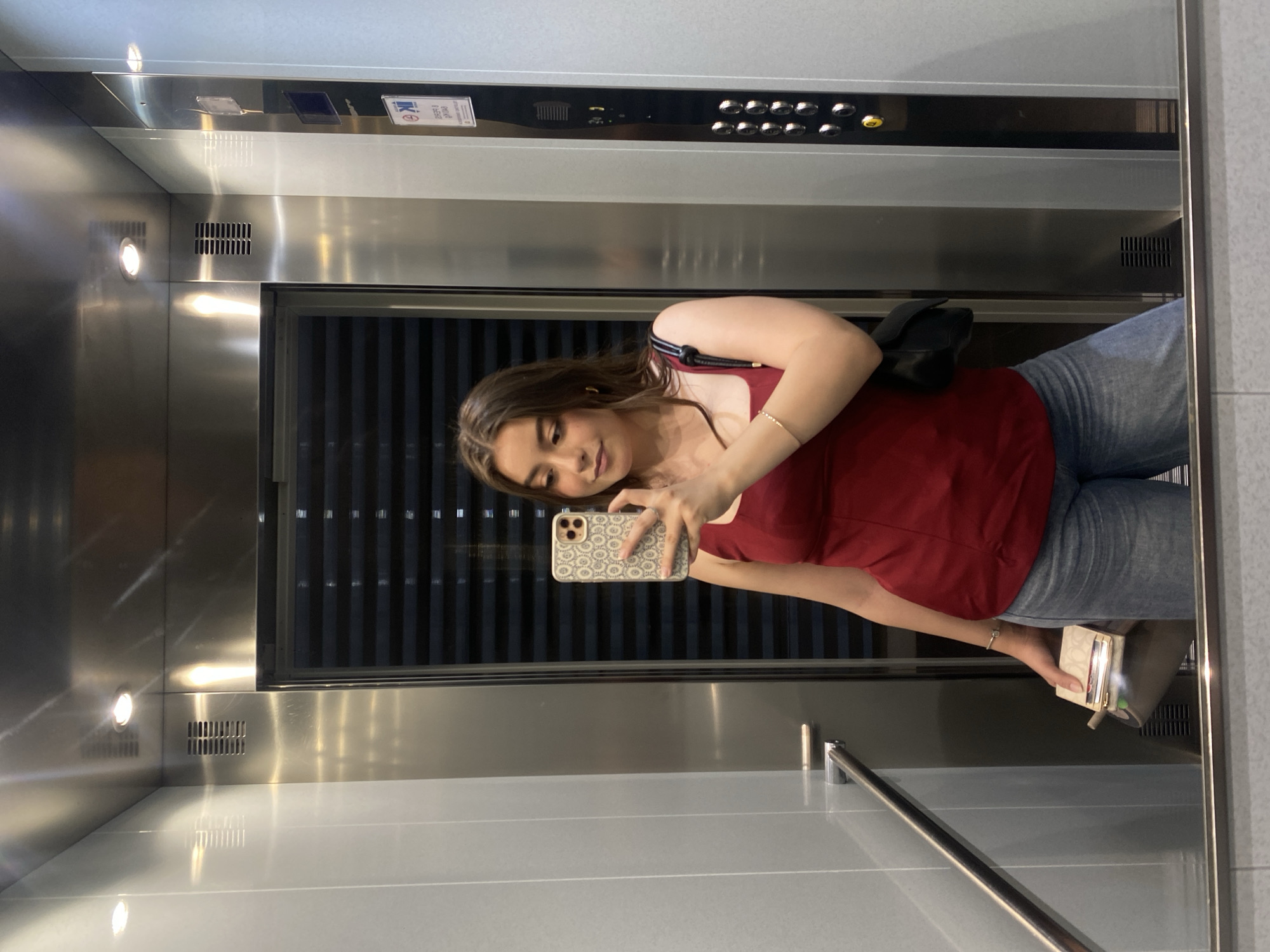 A young woman is taking a mirror selfie in an elevator. The elevator is stainless steel with a black panel containing buttons. The woman is wearing a red top and jeans, and she has a black purse over her shoulder. She is holding a phone in her left hand and a small, rectangular object (possibly a wallet or cardholder) in her right hand. The city and specific location are indeterminate.