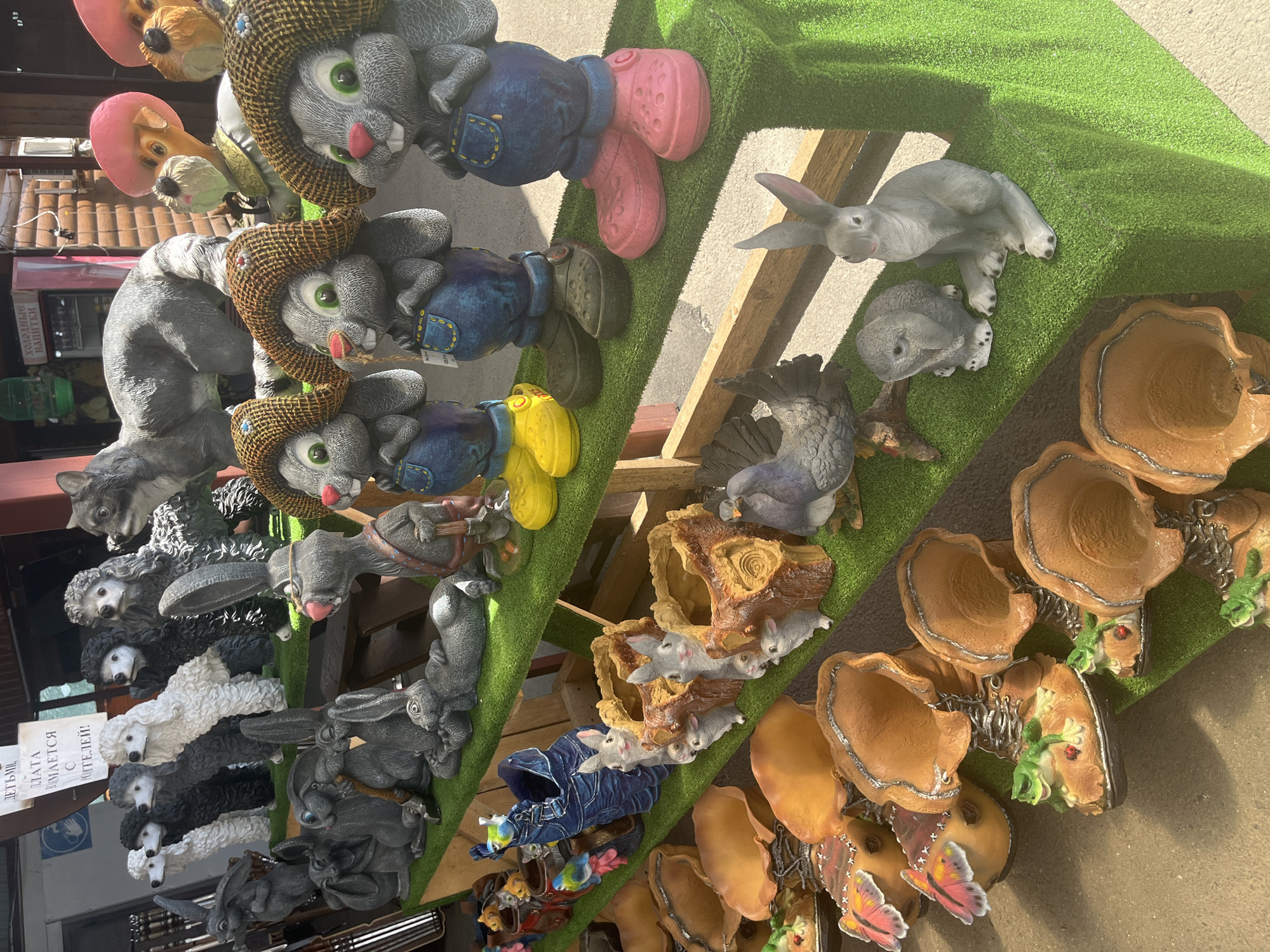 This image displays a market stall in Plastunovskaya, Russia, filled with an assortment of garden gnomes and statues. The items are arranged on tiered shelves covered with artificial green turf. On the top shelf, there are several grey, stylized dog figures, including poodles and a larger wolf-like statue. Further to the right are two tan dog-like statues wearing pink hats.
The middle shelves feature various rabbit figurines in overalls and hats, as well as a blue ceramic pot with a denim design adorned with small bird statues. There are also several brown, mushroom-shaped planters and a grey bird statue.
The bottom shelves showcase more of the brown, mushroom-shaped planters, some with attached frog figurines, and terracotta-colored pot-like items. In the foreground, several grey rabbit statues are visible, including one sitting upright and another with its head down. The overall scene suggests a retail environment selling decorative outdoor items.