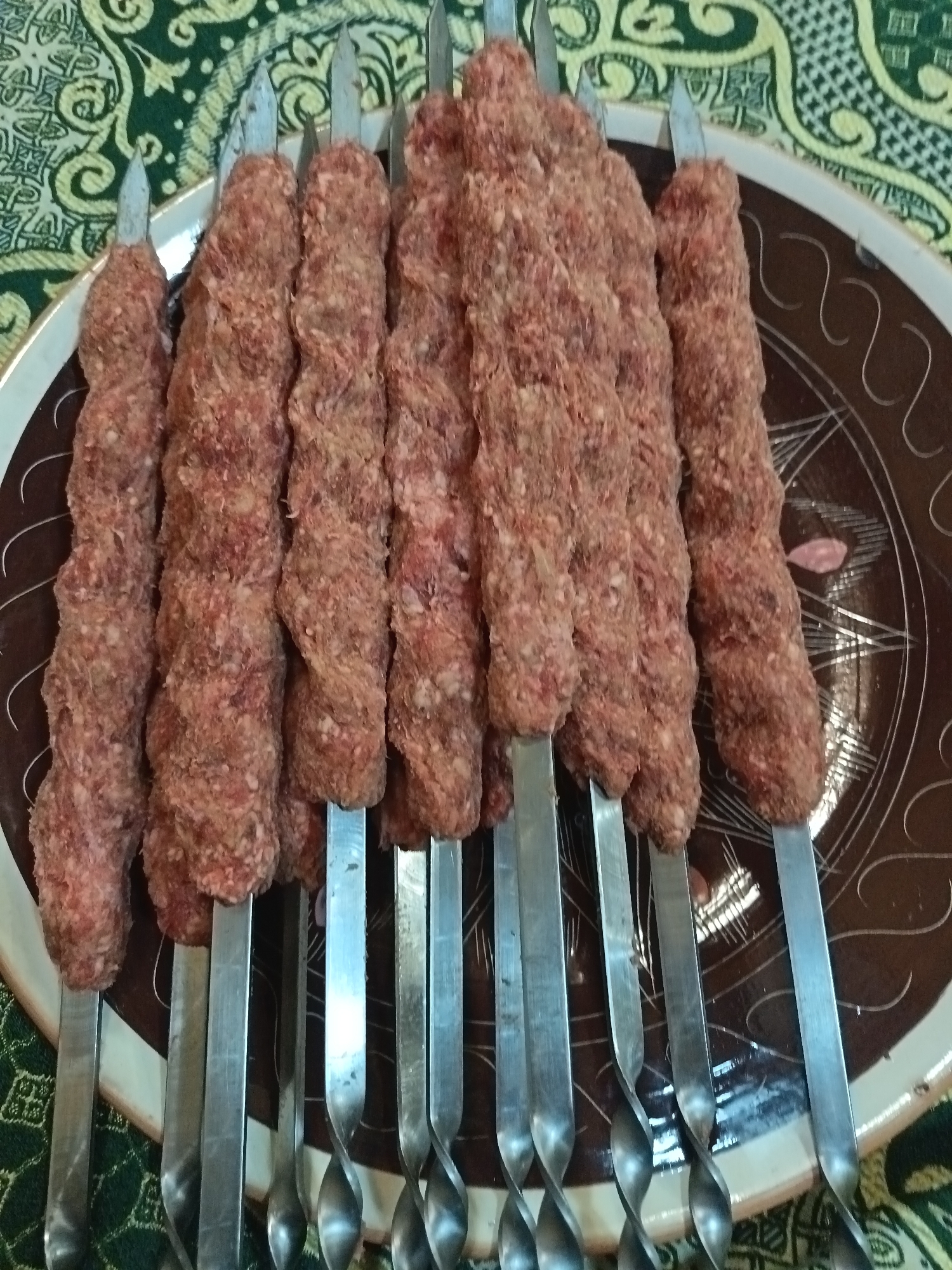 A platter of eight raw meat kebabs on metal skewers rests on a table covered with a green and gold patterned tablecloth. The kebabs are positioned closely together, aligned on the platter. The skewers are arranged in a vertical row. The platter is circular, brown, and displays a pattern of light-colored concentric circles and lines. The scene appears to be indoors. Nizhnevartovsk, Russia is not discernible from the image.