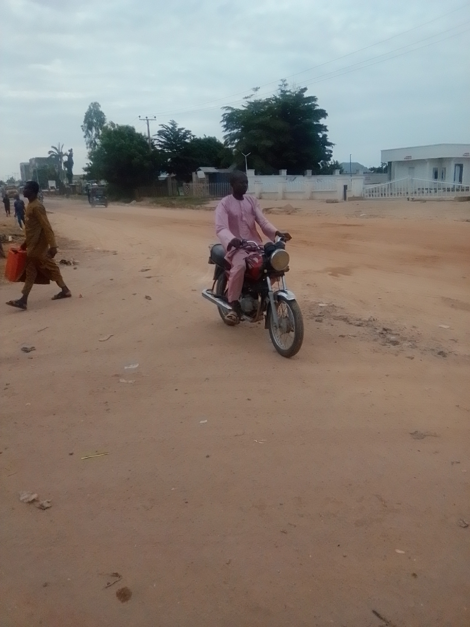 In Gombe, Nigeria, a dusty, unpaved road is the setting for everyday life. A man rides a motorcycle, his pink attire a stark contrast to the earthy tones of his surroundings. To the left, another man, dressed in a brown outfit, walks with a bright orange container, possibly filled with water or fuel. In the background, the scene is dotted with trees and buildings, including a white structure with a fence, hinting at the presence of homes or businesses. The sky is overcast, suggesting a mild climate. The overall impression is one of a typical day in a developing urban environment, where daily tasks are carried out amidst the natural and built landscape.