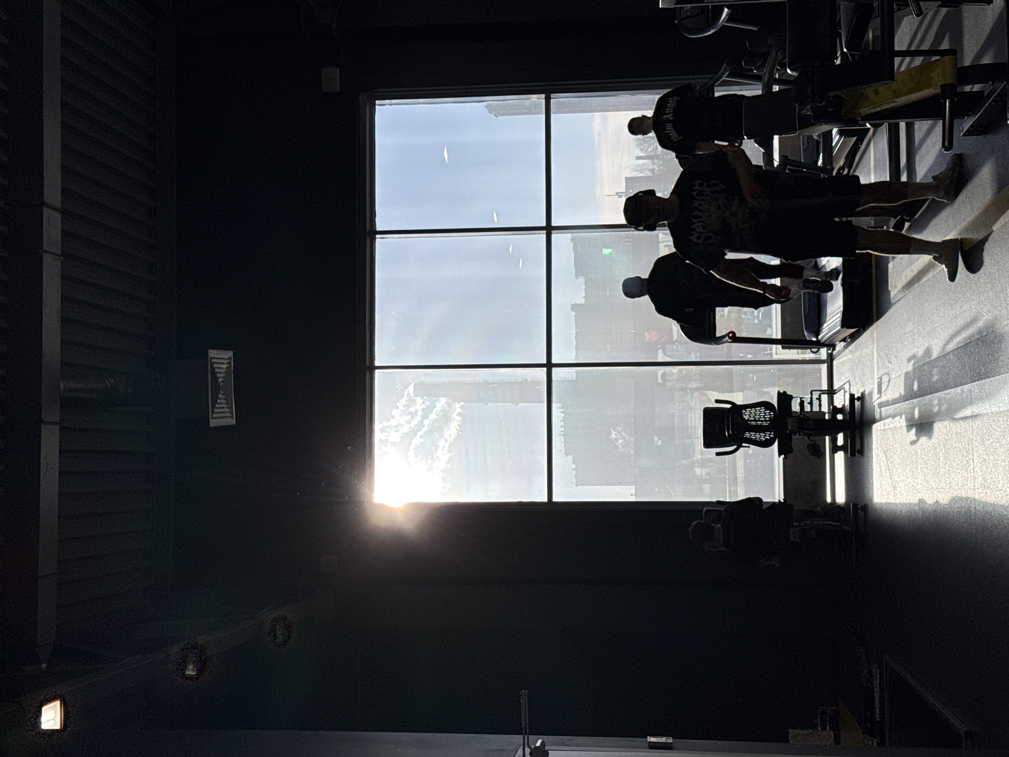 A dimly lit interior space, possibly a gym or fitness center, is dominated by a large window revealing an external view of the city of Saint Petersburg, Russia. Figures of people engaged in activities, potentially exercising, are silhouetted against the bright window. Exercise equipment is visible in the foreground, adding to the ambiance of a workout environment. The light from outside, possibly the sun, shines through the window, creating stark contrasts and silhouettes.