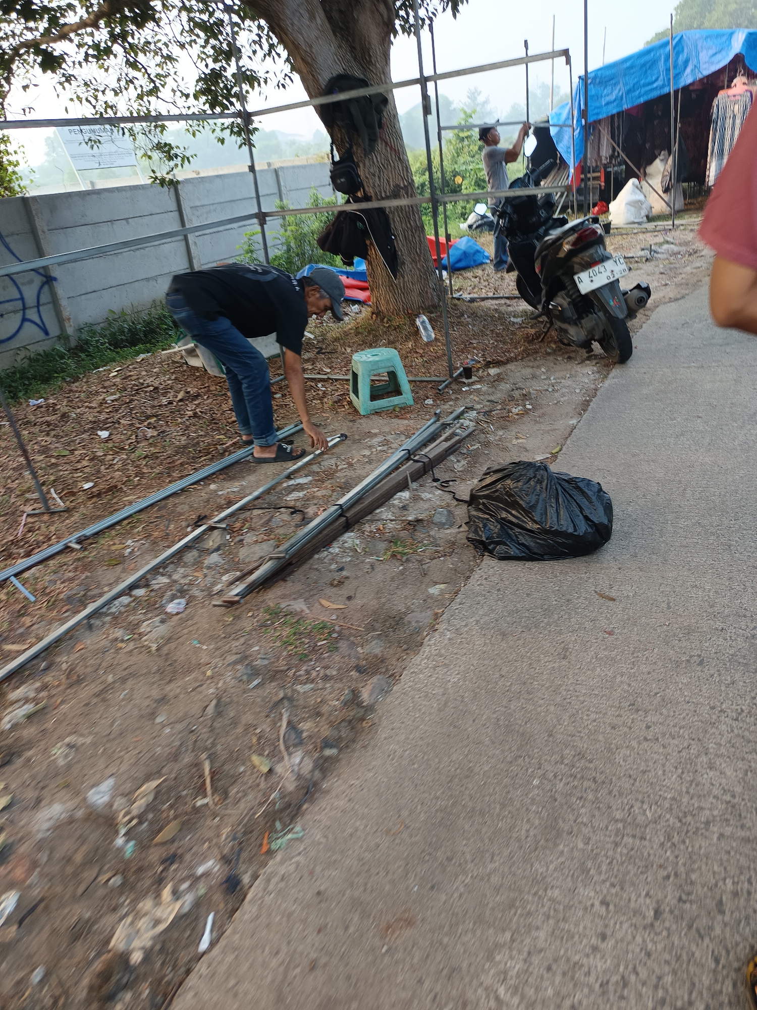 The image depicts an outdoor scene in the city of Sodong, Indonesia, with a focus on a man and construction materials. A man, wearing a black t-shirt, blue jeans, a gray cap, and sandals, is bent over and appears to be working with metal poles laid out on the ground. To his right, a black garbage bag is prominently placed on the paved surface. In the background, a motorcycle is parked, and a blue tarp covers a structure, suggesting a temporary setup or market stall. Various construction materials, including metal pipes and a green plastic stool, are scattered around the area. The environment is somewhat unkempt, with dry leaves and debris on the ground, and a concrete wall in the background. The overall impression is one of preparation or dismantling of a temporary structure in an urban outdoor setting.