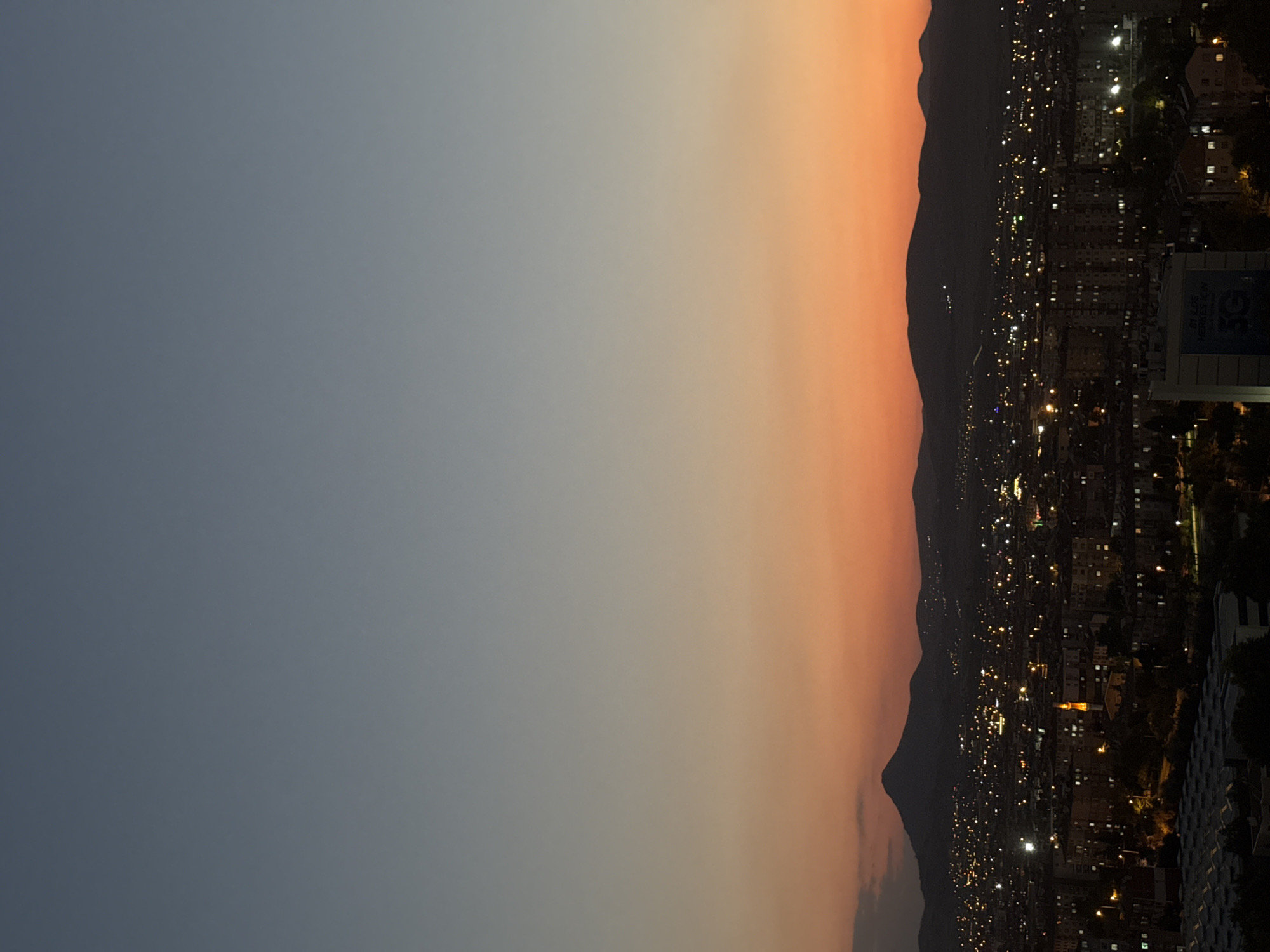 An aerial view captures the city of Konya, Turkey, at dusk. The sky transitions from a deep twilight blue at the top to a vibrant orange and pink gradient near the horizon, signaling the setting sun. Silhouetted against the colorful sky are rolling hills and mountains in the distance. Below, the urban landscape of Konya is illuminated by a multitude of lights from buildings and streetlamps, indicating a sprawling city. Residential buildings and apartment blocks are visible, interspersed with some taller structures. A prominent minaret, lit from within, stands out in the middle ground, suggesting the presence of a mosque. In the foreground, the tops of more buildings and possibly a large industrial or commercial structure with a patterned roof are visible. The overall impression is one of a bustling city settling into the evening under a dramatic sunset.