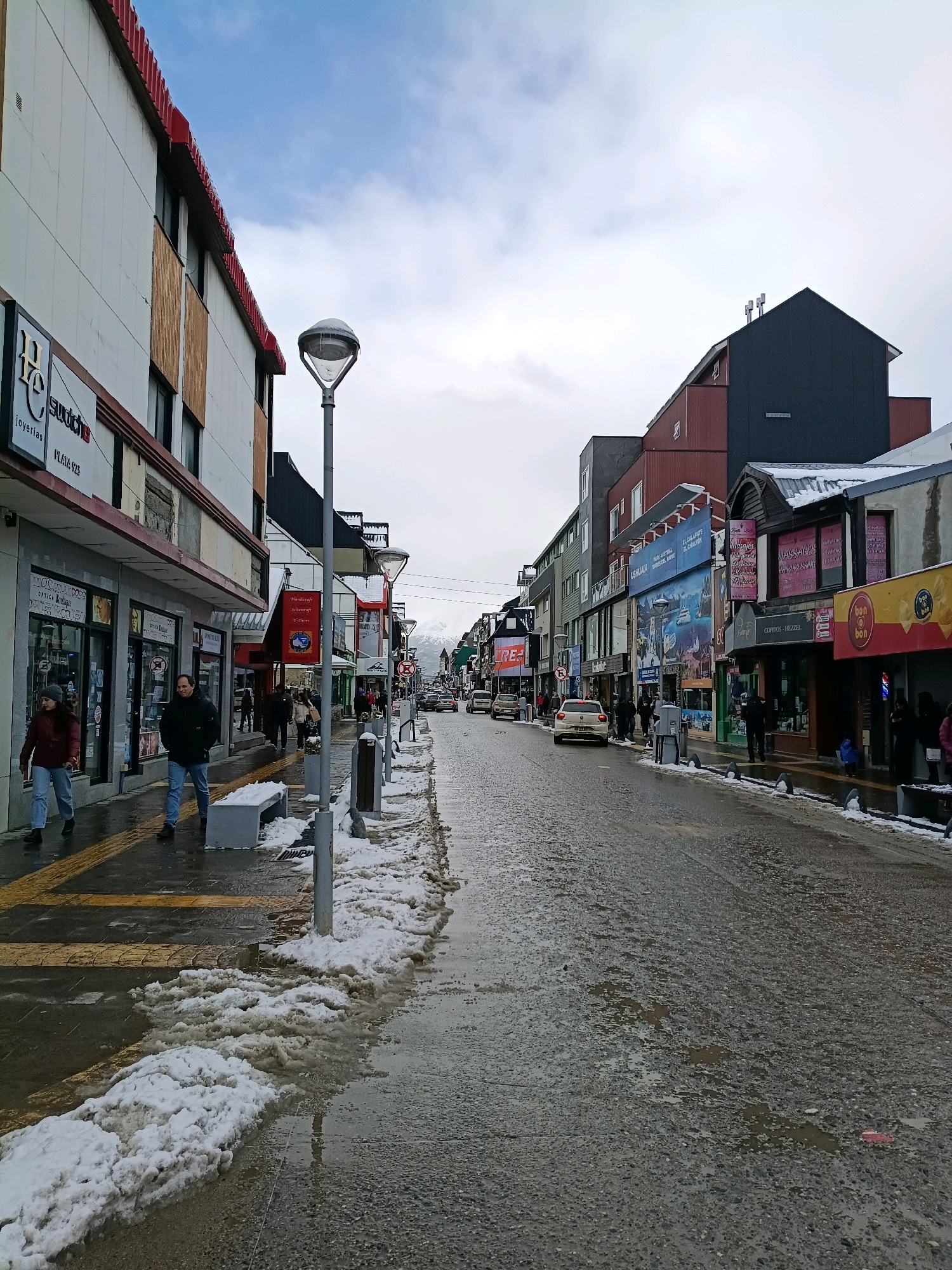 The image depicts a street scene in Ushuaia, Argentina, on a cloudy day with patchy snow and ice on the sidewalks and road. The street is lined with multi-story buildings housing various shops and businesses, indicated by signage such as "joyerías," "Swatch," "Óptica," "Handcraft," and "Bon o Bon." Several pedestrians are walking on the sidewalks, some wearing winter clothing like jackets and hats. A few cars are visible on the wet, icy road. Snowbanks and melting ice are present along the edge of the sidewalk and street. A street lamp with ice on its fixture stands on the sidewalk. The overall environment suggests a cold, wintery urban setting.