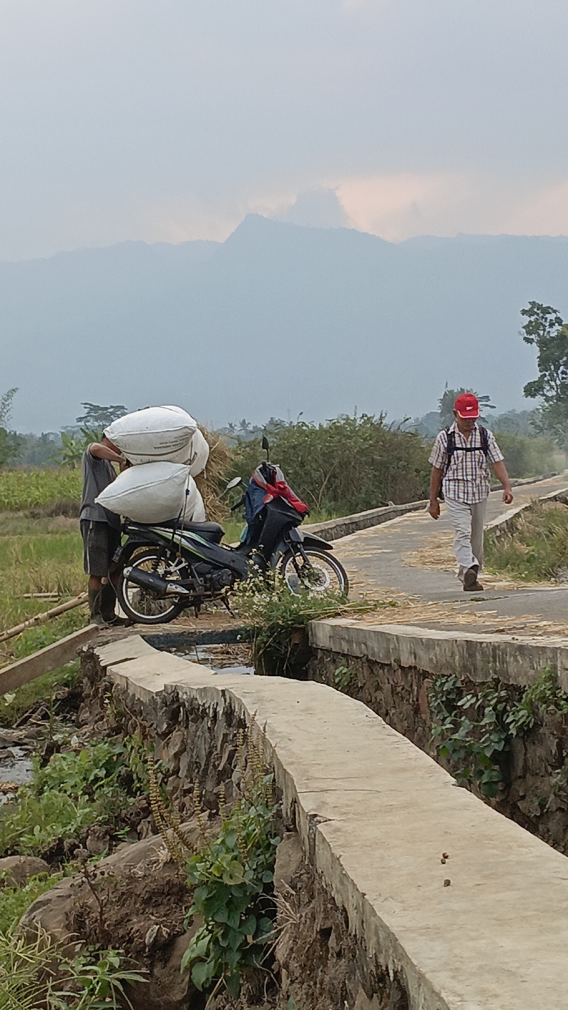 Two men are on a rural road in Salatiga, Indonesia, with a motorcycle. One man, on the left, wearing a gray t-shirt and shorts, is carrying two large white sacks on his shoulder. Next to him is a black motorcycle with two white sacks stacked on the seat and handlebars. The second man, on the right, is walking away from the motorcycle. He is wearing a red cap, a checkered shirt, light-colored pants, and dark shoes, with a backpack on his shoulders. The road appears to be a bridge or raised path, with a water channel below. The background shows agricultural fields, lush green vegetation, and hazy mountains in the distance under an overcast sky.