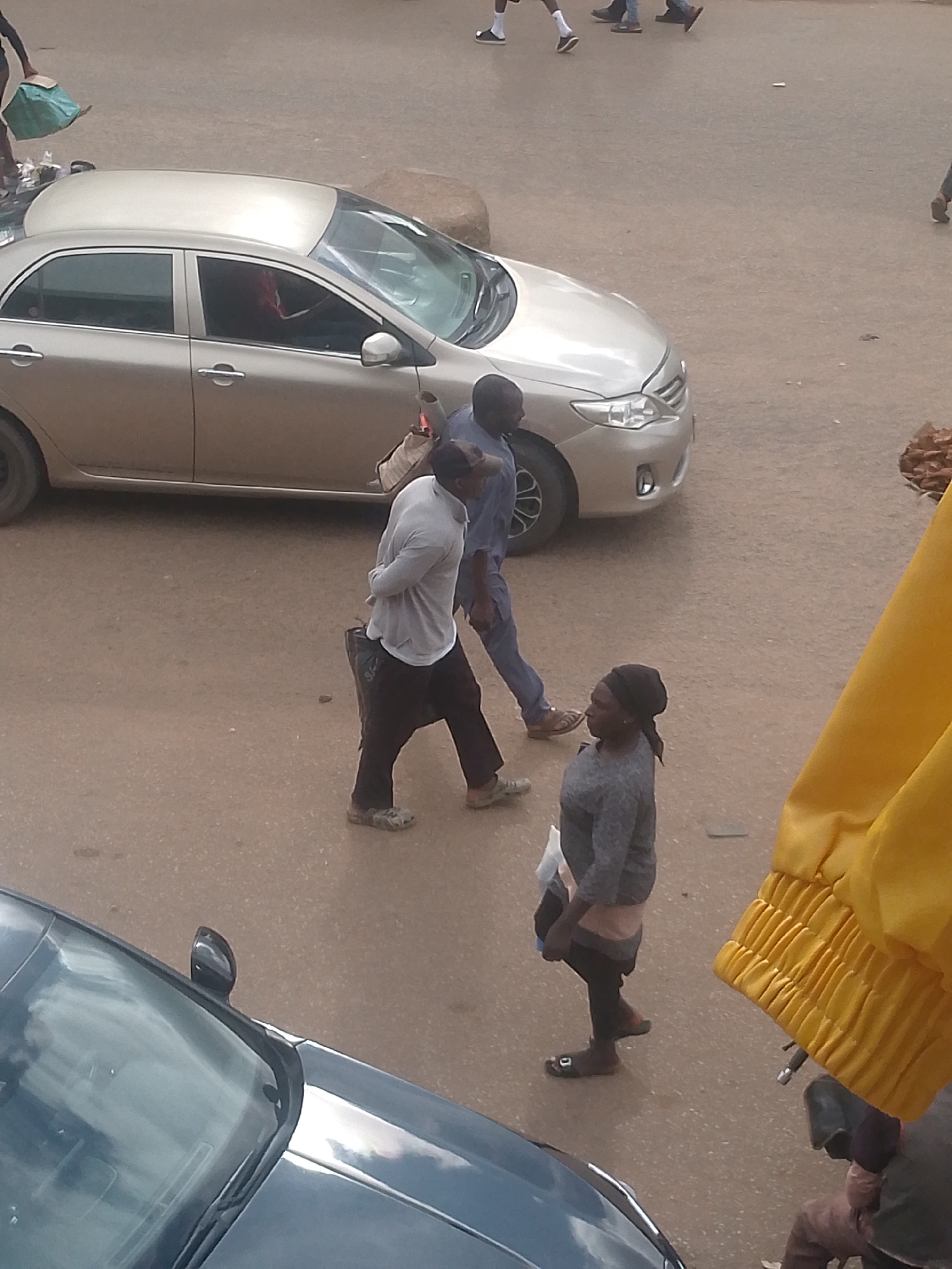 The image depicts a street scene in Bukuru, Nigeria. In the foreground, a silver sedan is parked on the asphalt road. Several people are present. To the left of the sedan, a person is walking away from the camera, carrying a green bag. To the right of the sedan, two individuals are walking towards the right side of the frame. One is wearing a light-colored long-sleeved shirt and dark pants, and appears to be carrying a dark bag. The other is wearing a blue shirt and dark pants, and has a bag slung over their shoulder. In the lower right corner, a woman wearing a grey sweater and black leggings is standing, looking to her left. A bright yellow fabric is visible in the far right of the image, partially obscuring a motorcycle or scooter. The overall environment appears to be an outdoor market or bustling street.