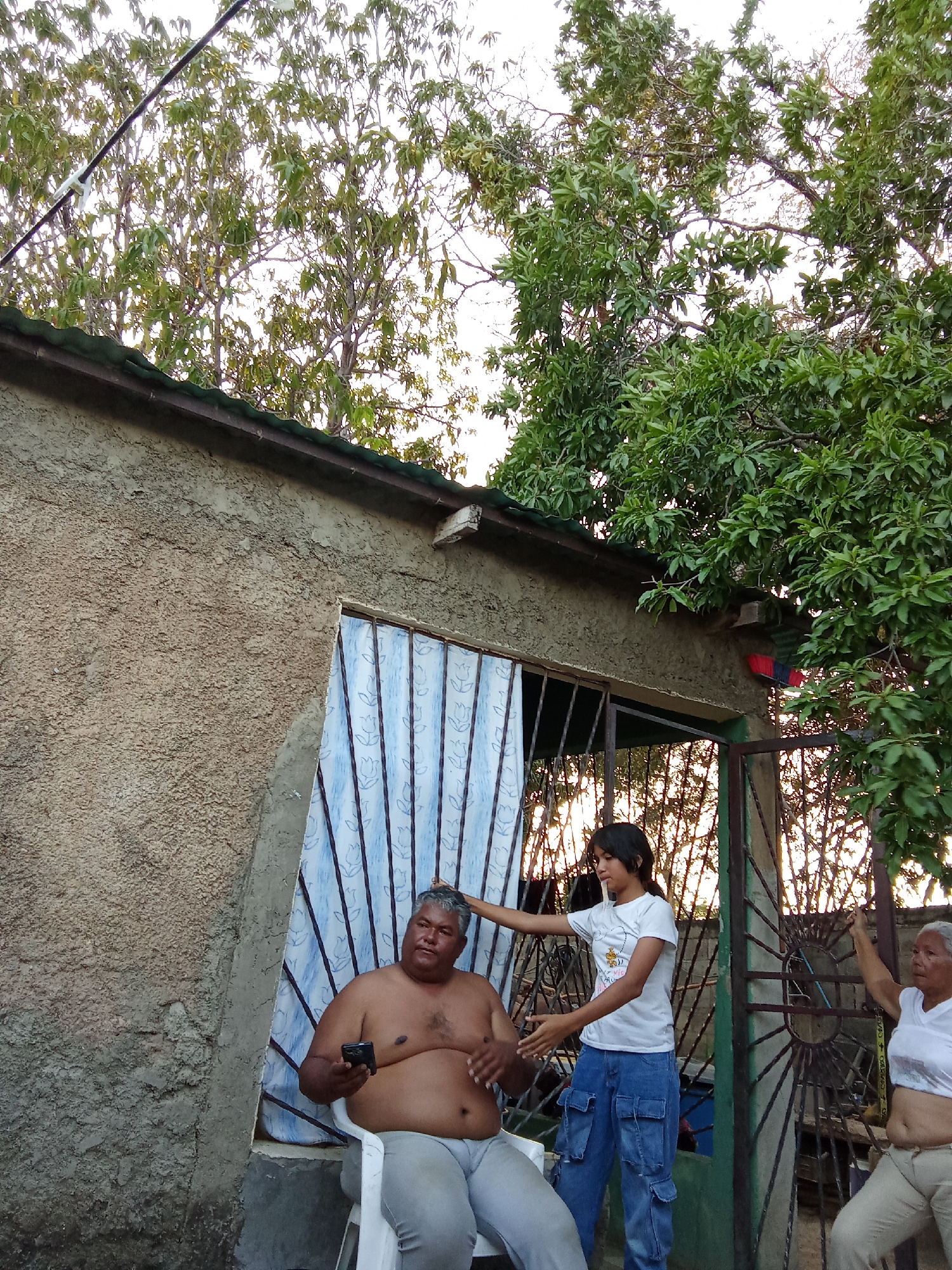 A residential scene in Juan Griego, Venezuela, shows a shirtless, overweight man sitting in a white plastic chair outside a stucco-walled building with a green corrugated metal roof. The man holds a smartphone in his left hand. A young woman wearing a white t-shirt and blue cargo pants stands next to him, her right hand touching his head. An older woman in a white t-shirt stands to the right, holding onto a green metal gate featuring a circular design. The gate leads to an outdoor area with trees visible behind it. A red-and-blue object is attached to the wall above the gate. The building features a window with patterned blue curtains and a metal grate. Trees are visible above the building. The time of day is likely evening.