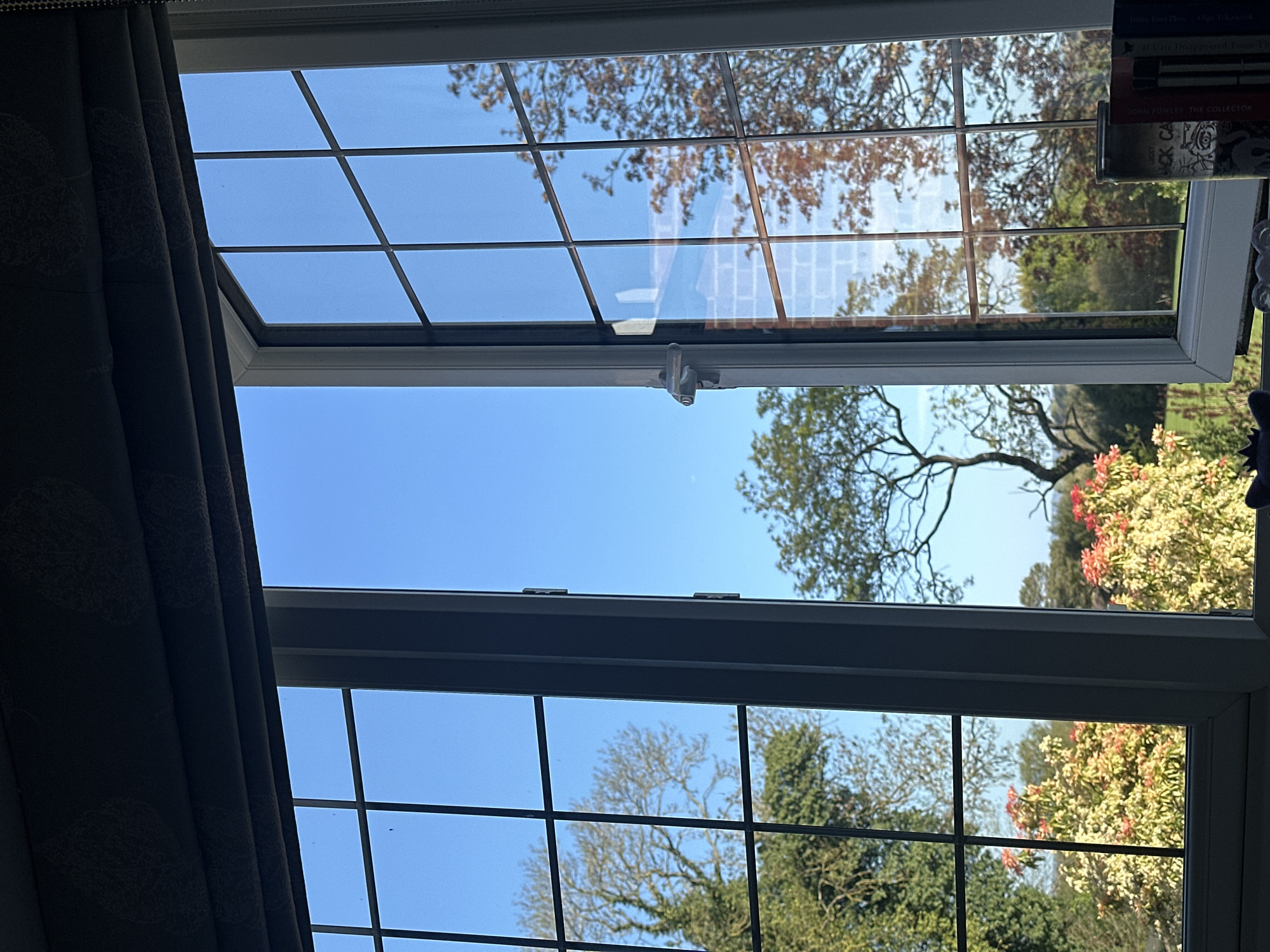 The image shows a view through an open window with multiple panes. Outside, there are trees and flowering bushes, suggesting a garden or park setting. The sky is blue and clear. Inside, a dark curtain is visible on the left side of the window. On the right, there are books on a shelf. The location is likely Wealden, United Kingdom. The specific content of the books on the shelf include "Drive Your Flow" by Diya Tokarczuk, "If Cats Disappeared from the World" and "John Fowles The Collector." The lighting suggests it is daytime.