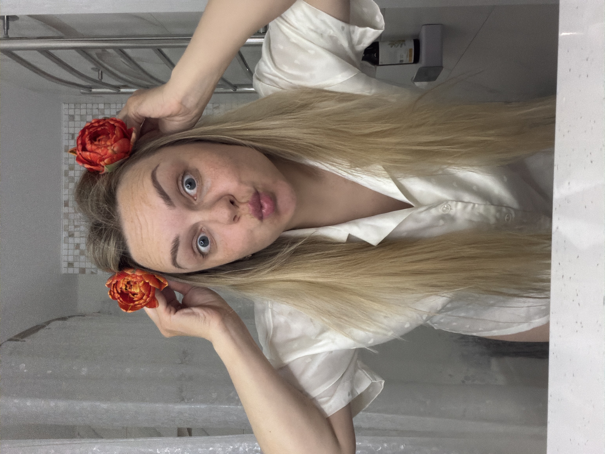 A woman with long blonde hair is in a bathroom setting. She holds two orange flowers, one near each side of her head. She is wearing a white shirt and appears to be making a facial expression with pursed lips. The bathroom features white and grey tiles, a shower curtain, and a towel rack. A bottle is visible on a shelf in the background. The location and details do not inherently indicate the city of Votkinsk, Russia.