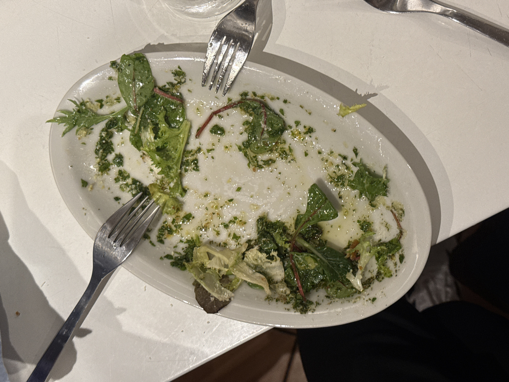 A close-up shot shows a partially consumed salad on an oval-shaped white plate, with two forks resting on the plate and table. The salad consists of leafy greens, possibly lettuce and spinach, and is dressed with a green sauce, potentially pesto. The plate is set on a white table, and the edge of a drinking glass is visible in the top center of the frame. Clothing suggests someone is seated at the table. The exact location in Belgrade, Serbia, is unknown.
