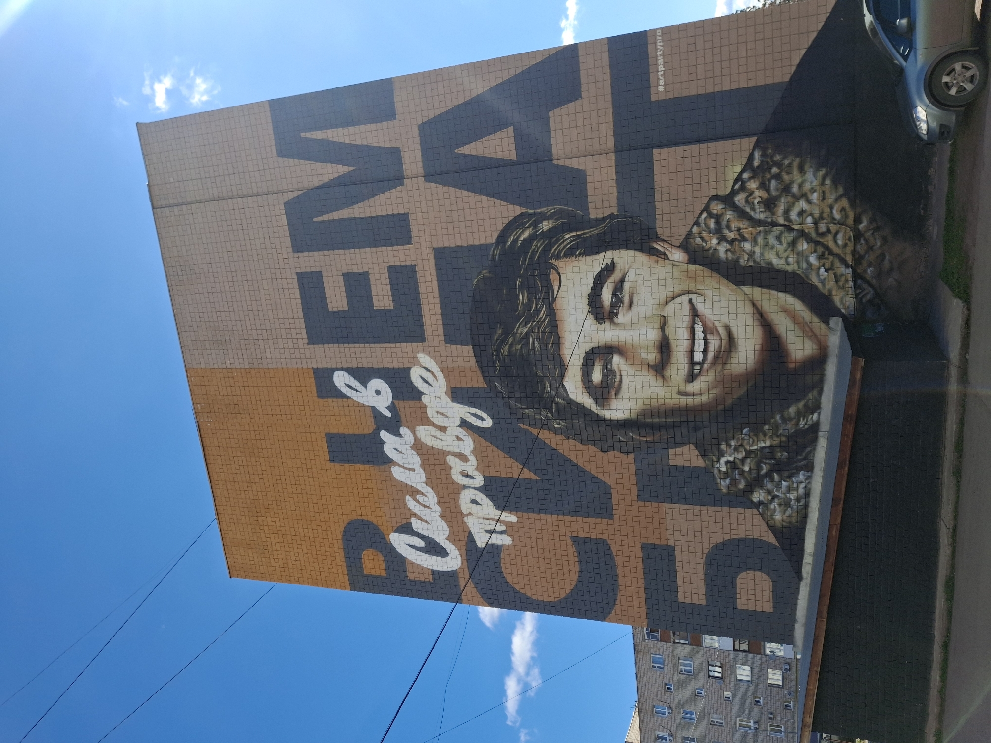 A low-angle shot of a mural in Sarapul, Russia, displays a smiling man's face superimposed over blocky lettering. The mural is painted on the side of a brick building. The lettering appears to be in Cyrillic. Below the face, there is Cyrillic text in white lettering. A car is parked alongside the building. Overhead power lines and a bright blue sky with a few clouds are visible in the background. Another building with multiple windows is visible on the right side of the image.
