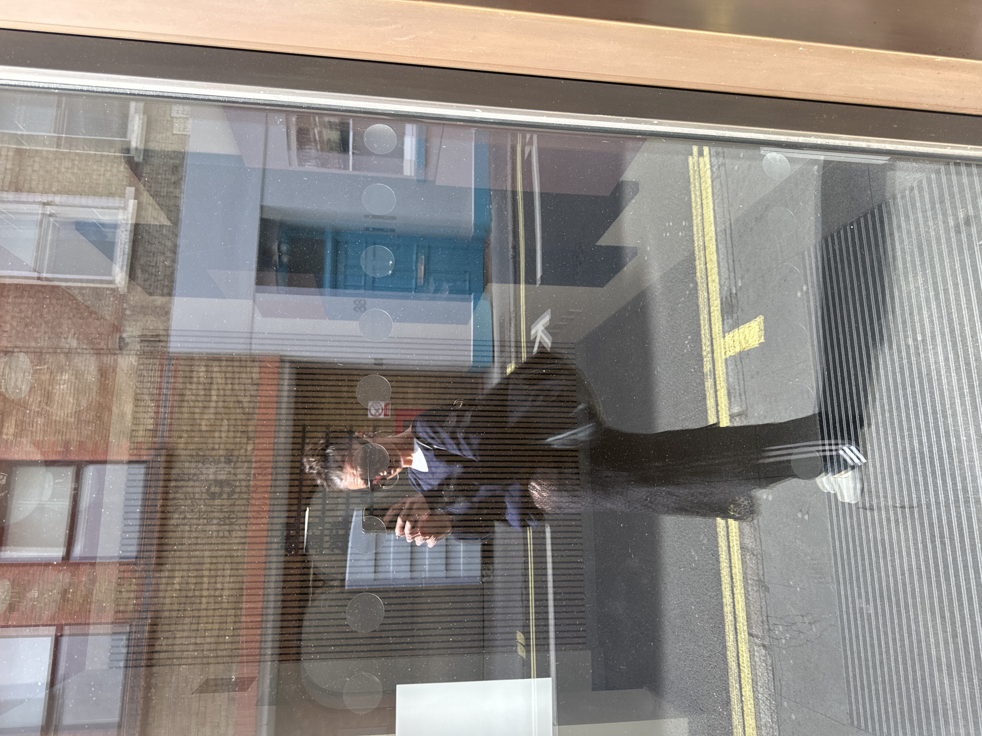 A mirrored reflection shows a person holding a phone in a storefront window. The reflection also includes part of a brick building with windows, a blue building with a door, and a street with yellow lines. The person is wearing a dark shirt and dark pants with light-colored stripes on the side. The reflection suggests a city location, likely in London, United Kingdom, given the architectural styles and street markings typical of the area. The setting is urban, with a mix of residential and commercial buildings visible in the reflection.