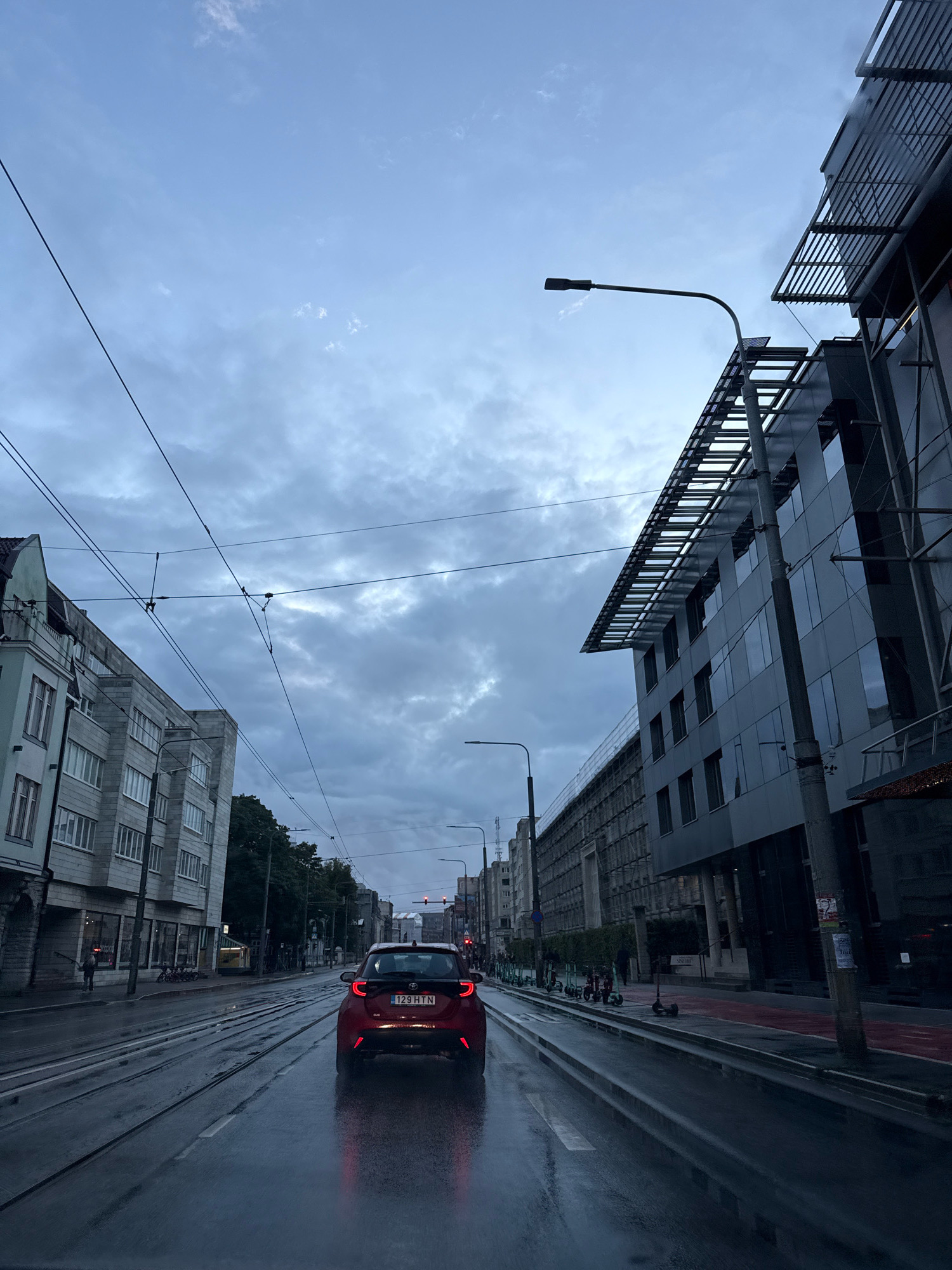 This is a ground-level, rear-facing medium shot captured on a wet street in Tallinn, Estonia. The overcast sky is a dominant feature, with a mix of light and dark grey clouds. A red car, identified by license plate 129 HTN, is in the foreground, driving away from the camera and reflecting the red brake lights onto the wet asphalt. The street is lined with tram tracks.
To the left of the street, a multi-story building with a light-colored facade and many windows is visible. Further down the street on the left, more buildings and trees can be seen. To the right of the street, a modern, dark-colored building with a reflective facade and a slanted roof structure is prominent. A lamppost extends from the right side of the frame, angled towards the sky. Several electric scooters are parked in a line along the sidewalk on the right side of the street. Overhead, complex arrays of power lines crisscross the sky, suggesting the presence of public transportation like trams. The overall atmosphere is that of a damp, urban environment.
