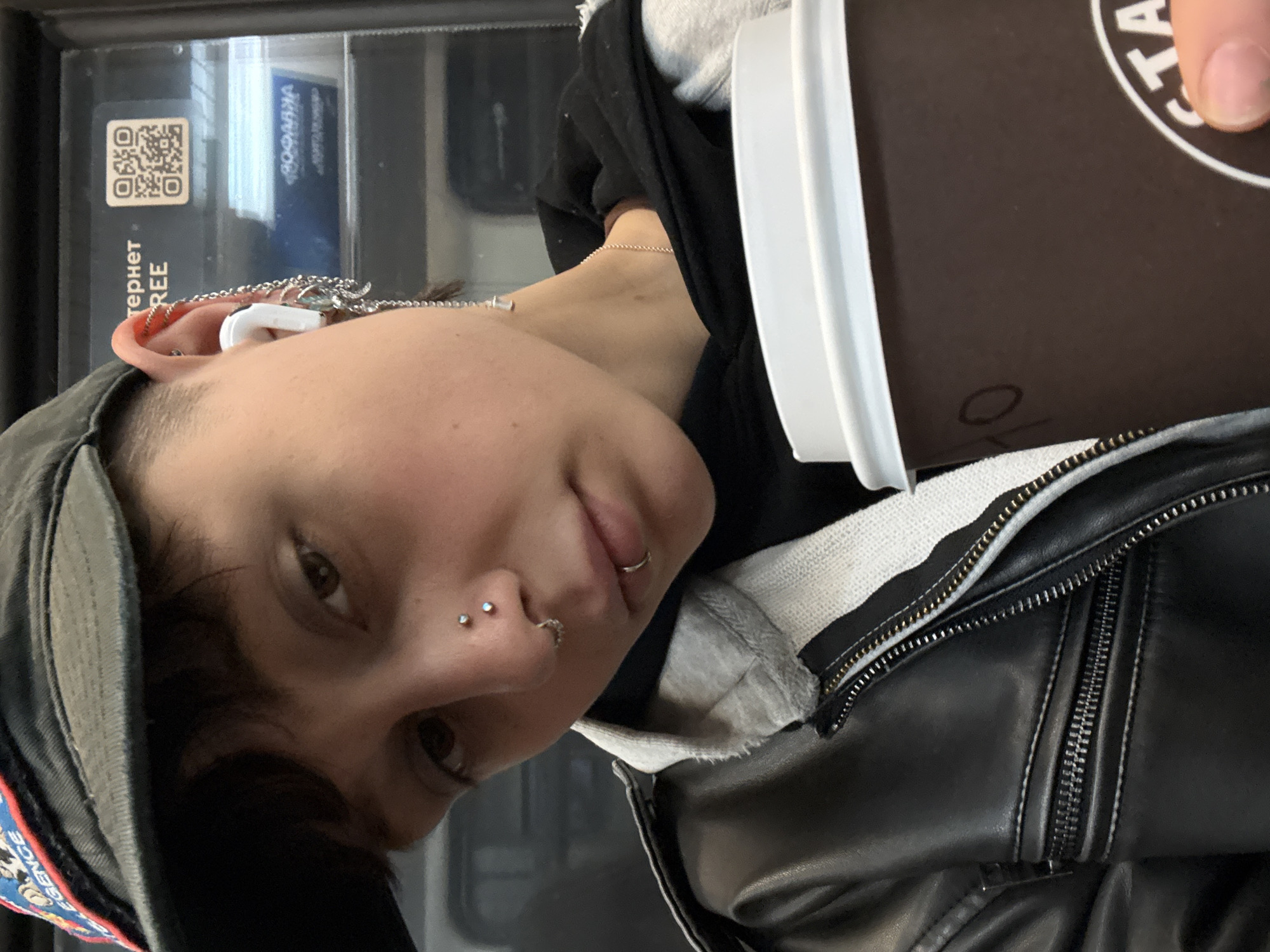 A person is holding a coffee cup while riding public transportation. The person is wearing a baseball cap, a black leather jacket, and a gray hooded sweatshirt. The person has multiple piercings on their nose and lip, and is wearing an earbud in their left ear. The background appears to be the interior of a train or bus. A sticker with a QR code and Russian text indicates there is free internet, which is on the Moscow Metro.